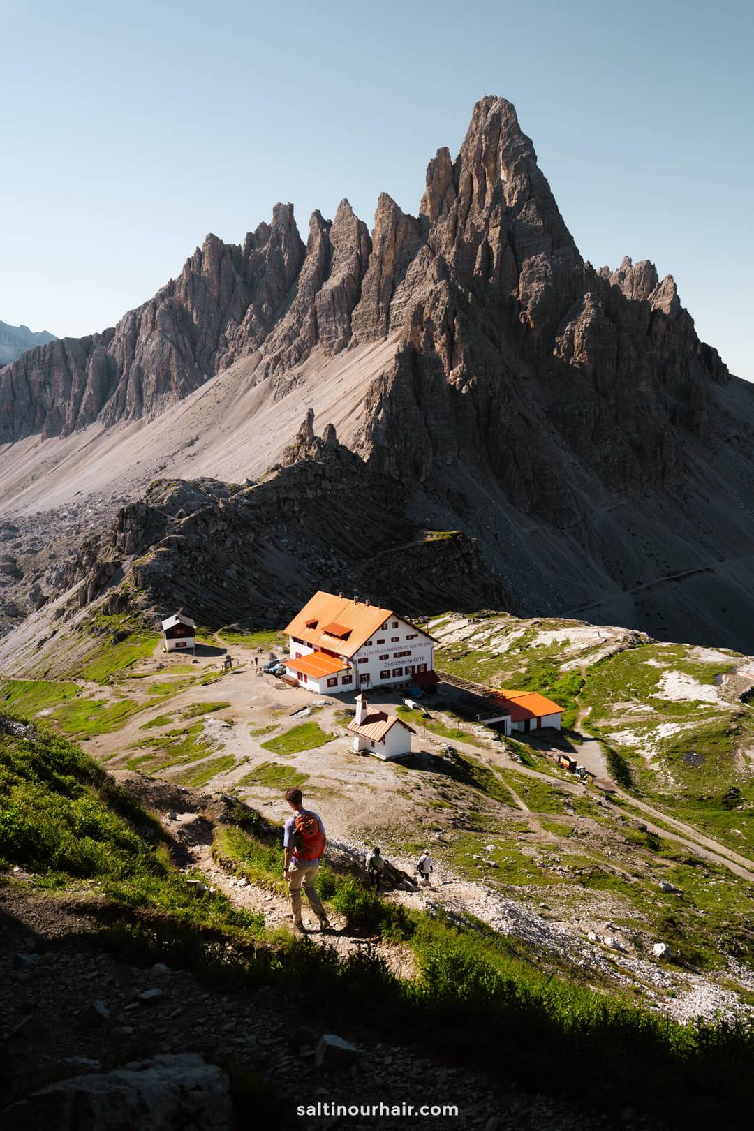 A person hikes uphill on a trail toward a group of alpine buildings, with the iconic Tre Cime&rsquo;s jagged mountain peaks rising in the background under a clear sky.