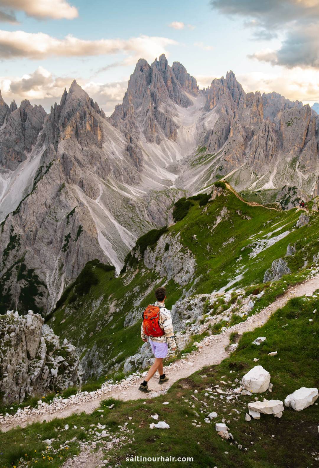 A person with a red backpack walks along a winding mountain trail beneath the iconic Tre Cime, surrounded by rocky peaks and green slopes under a partly cloudy sky.