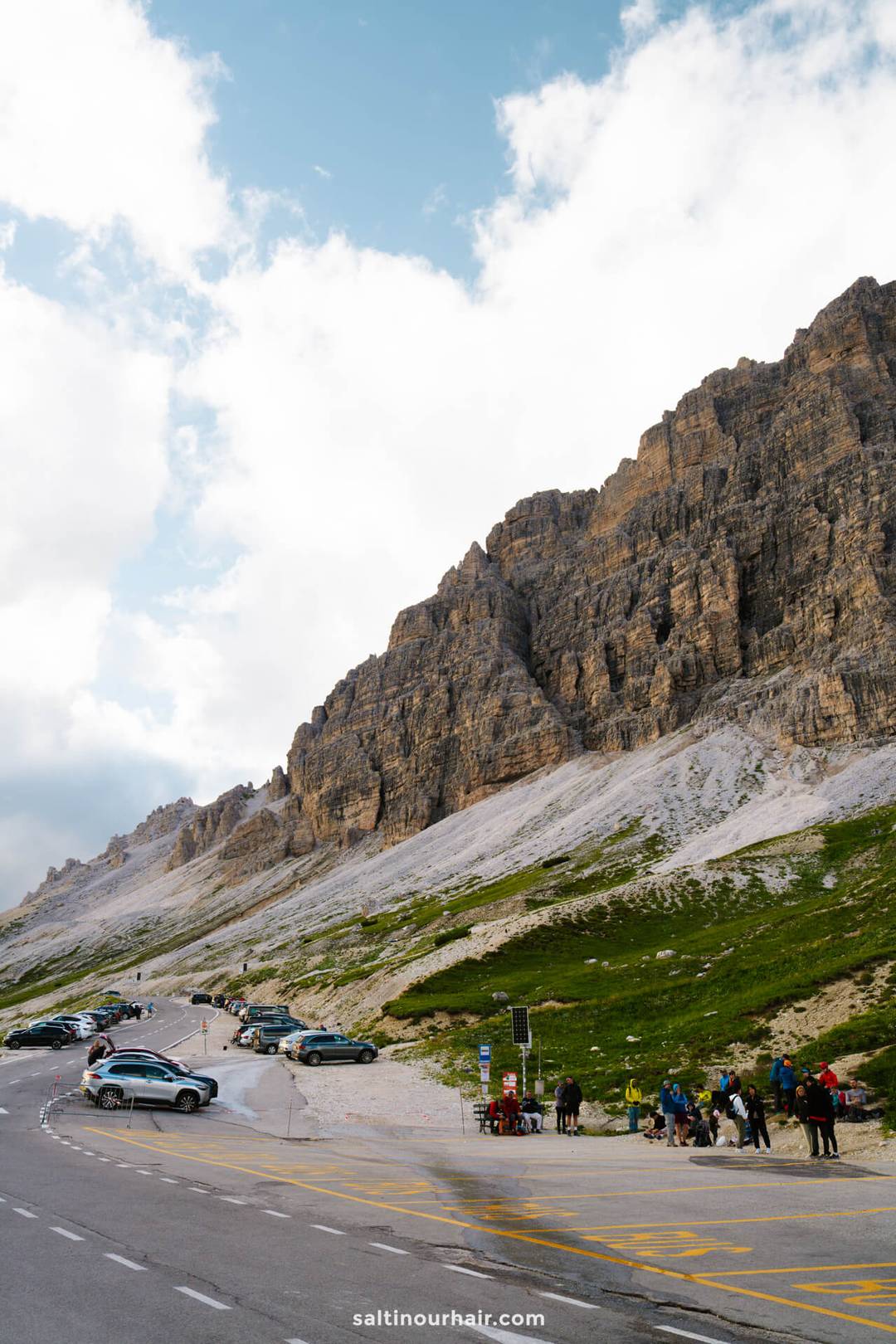 A mountain road with parked cars curves beside the steep rocky cliffs of Tre Cime. People gather near the roadside, and clouds fill the blue sky overhead.