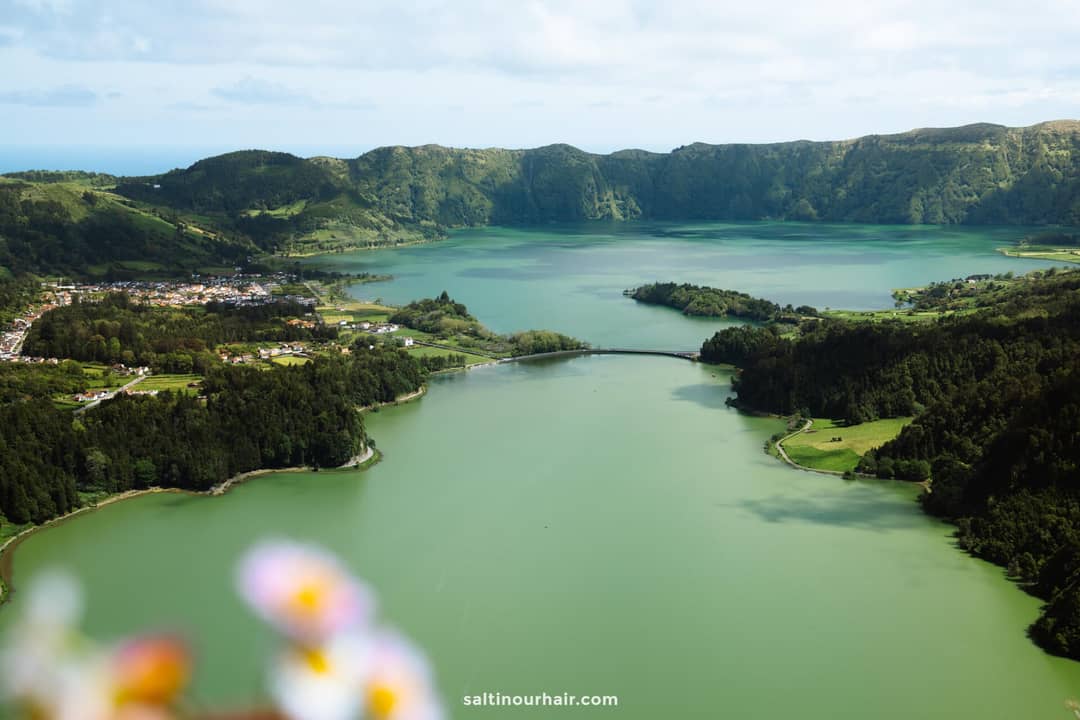 Luchtfoto van het groene meer van Sete Cidades omringd door weelderige heuvels, met een klein stadje en een brug over het water.
