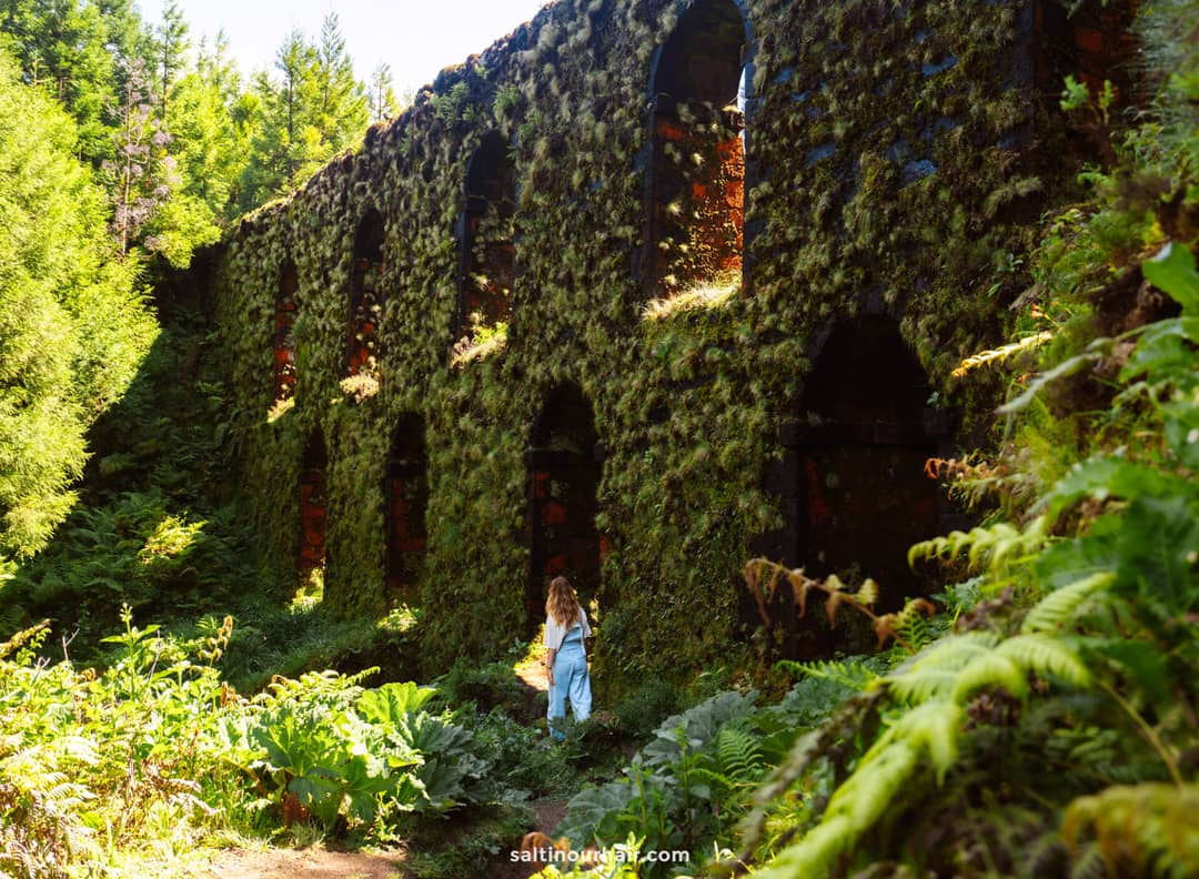 Een vrouw staat bij Muro das Nove Janelas, bedekt met mos en vegetatie, omringd door weelderig groen en zonlicht in het betoverende landschap van Sete Cidades, Sao Miguel Azoren