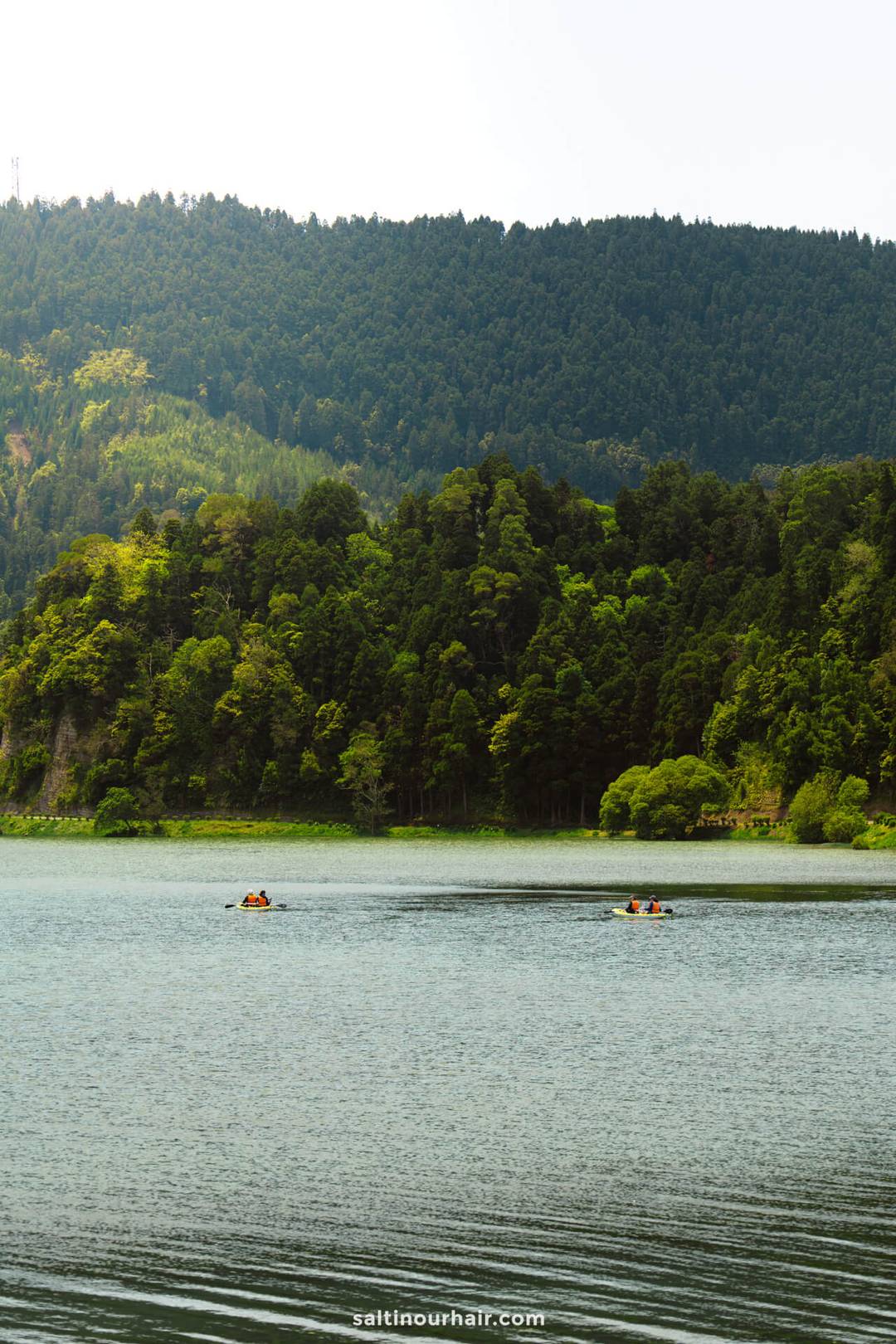 Twee kajaks met mensen peddelen op een rustig meer omringd door dicht, groen bos en heuvels onder een bewolkte hemel &mdash; een van de leukste dingen om te doen in S&atilde;o Miguel.