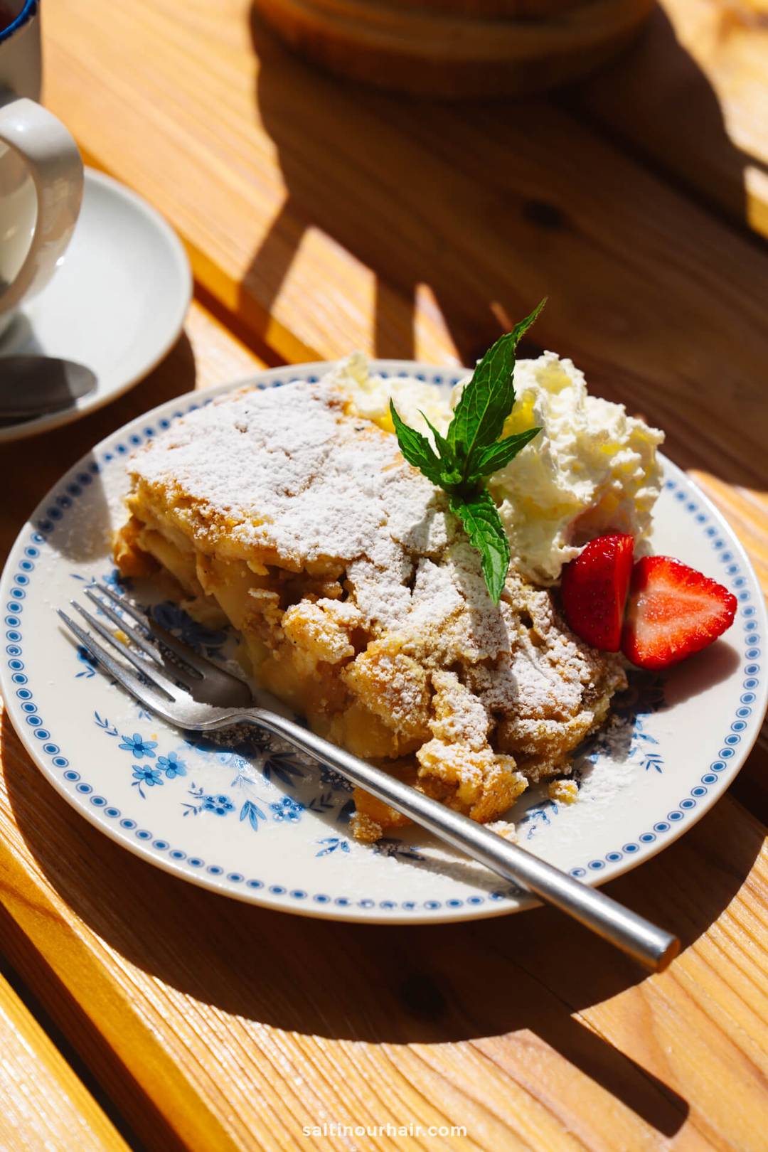 A plate with a slice of apple crumble topped with powdered sugar, whipped cream, strawberries, and a mint sprig sits on a wooden table with a fork&mdash;perfect for savoring after a hike on the scenic Alpe di Siusi.