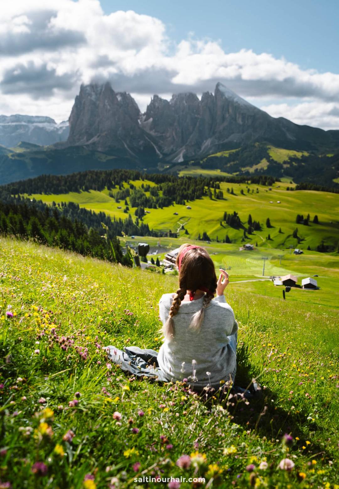 A woman with braided hair sits in a grassy field of wildflowers on the Seiser Alm, facing distant Dolomites mountains and a valley dotted with houses under a partly cloudy sky in Italy.