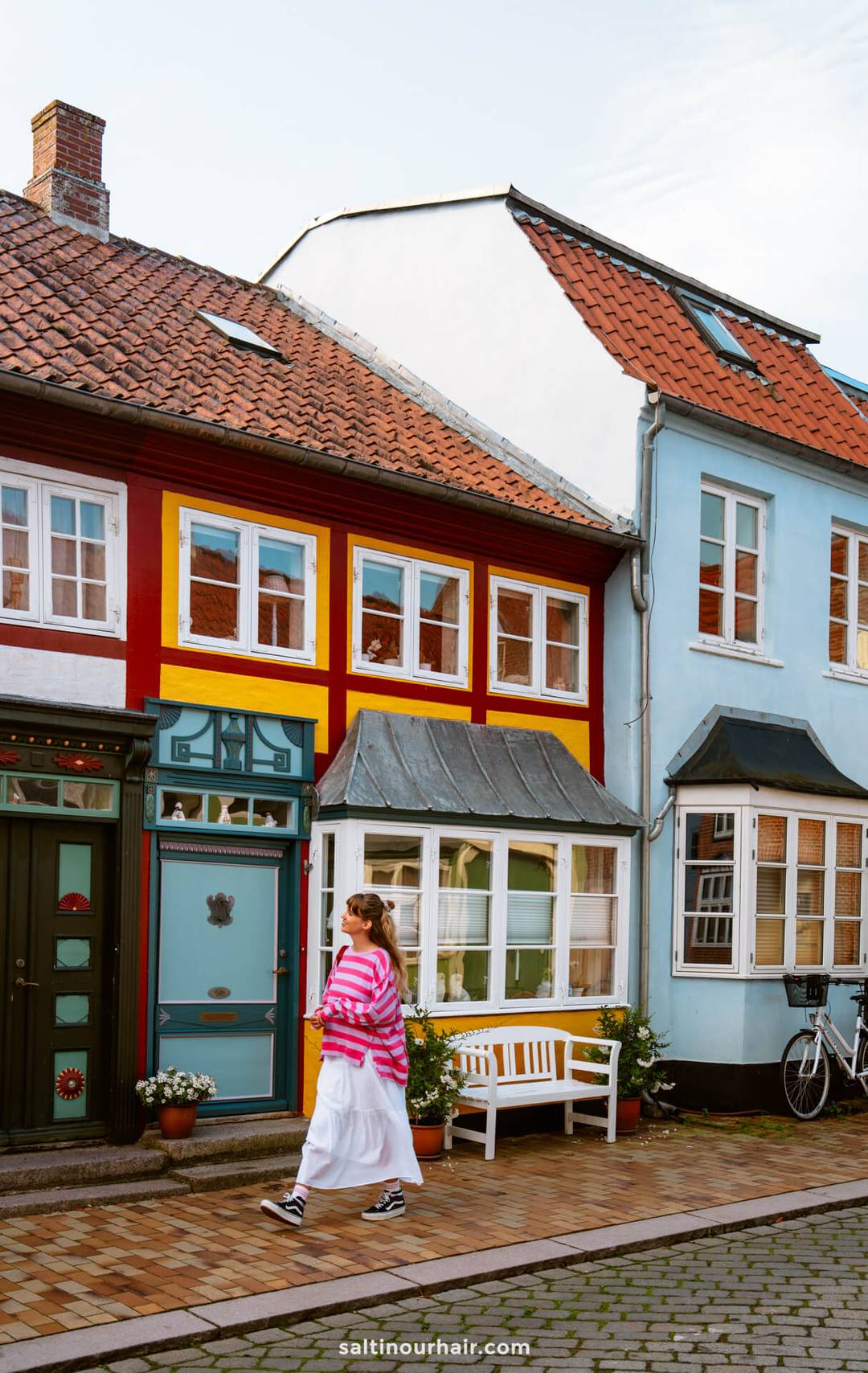 A person wearing a pink striped top and white skirt walks past colorful houses with red, yellow, and blue facades on a cobblestone street&mdash;perfect inspiration for your Denmark itinerary.