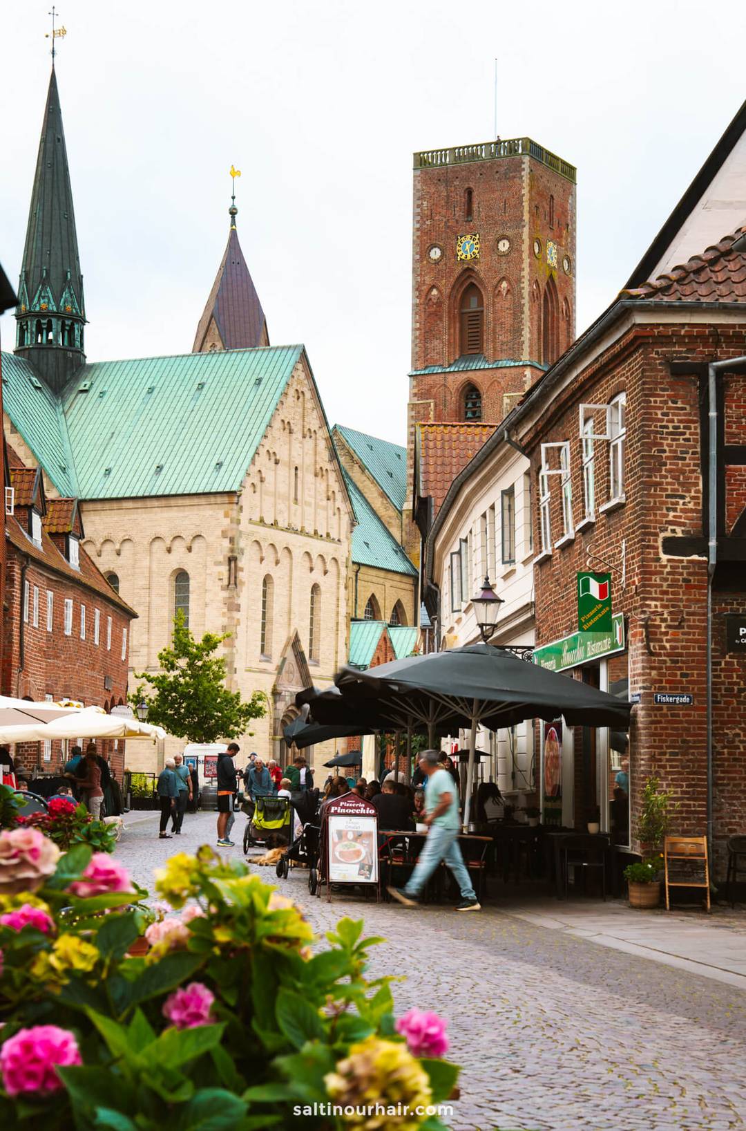 A cobblestone street with outdoor cafes and pedestrians, lined with brick buildings, leads to a large historic church with a tall tower and green roof in Ribe&mdash;an ideal sight on any Denmark itinerary.