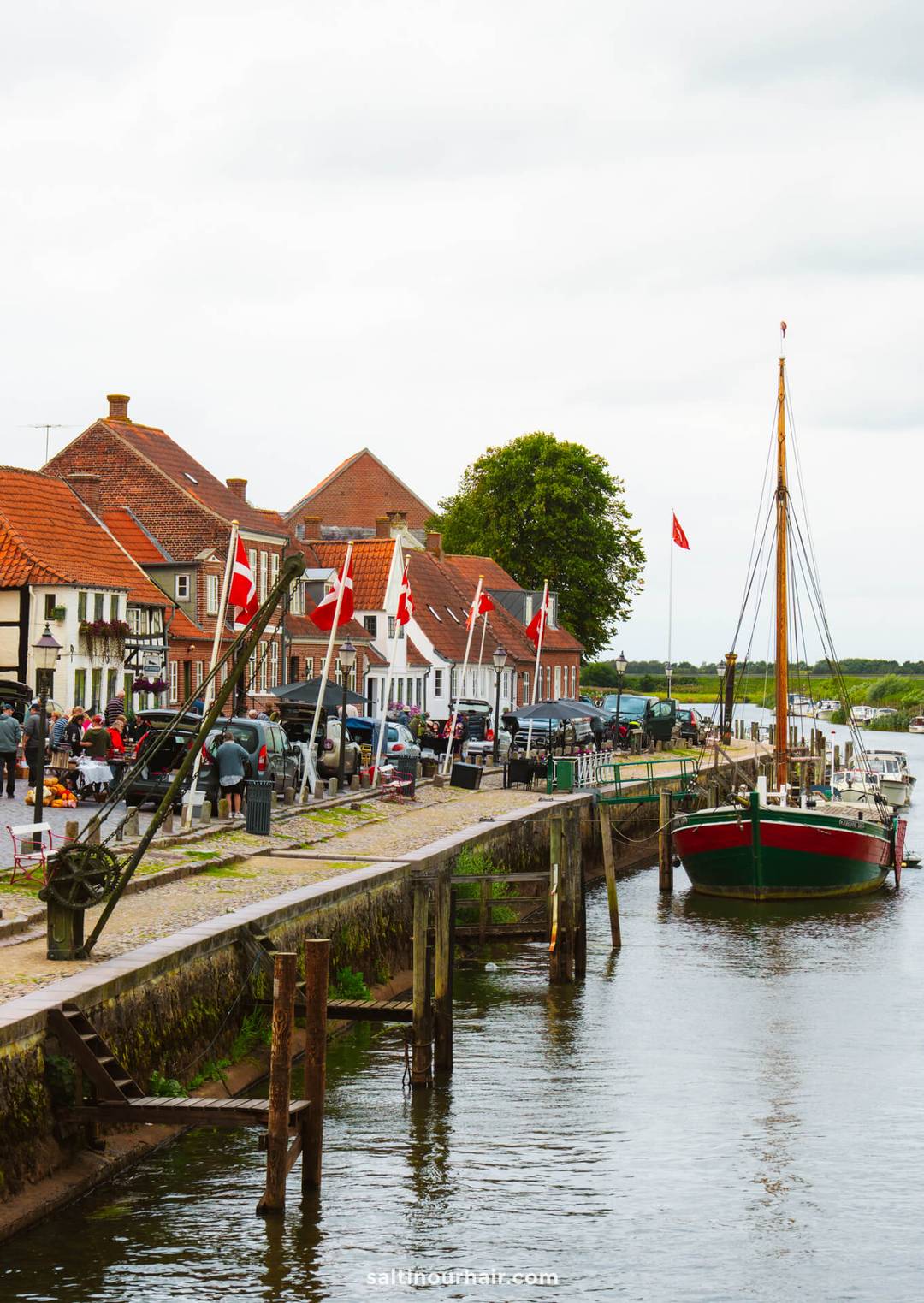 A docked sailboat on a canal beside a walkway lined with red-roofed buildings in Ribe, with several red-and-white flags. People are gathered along the scenic waterfront.