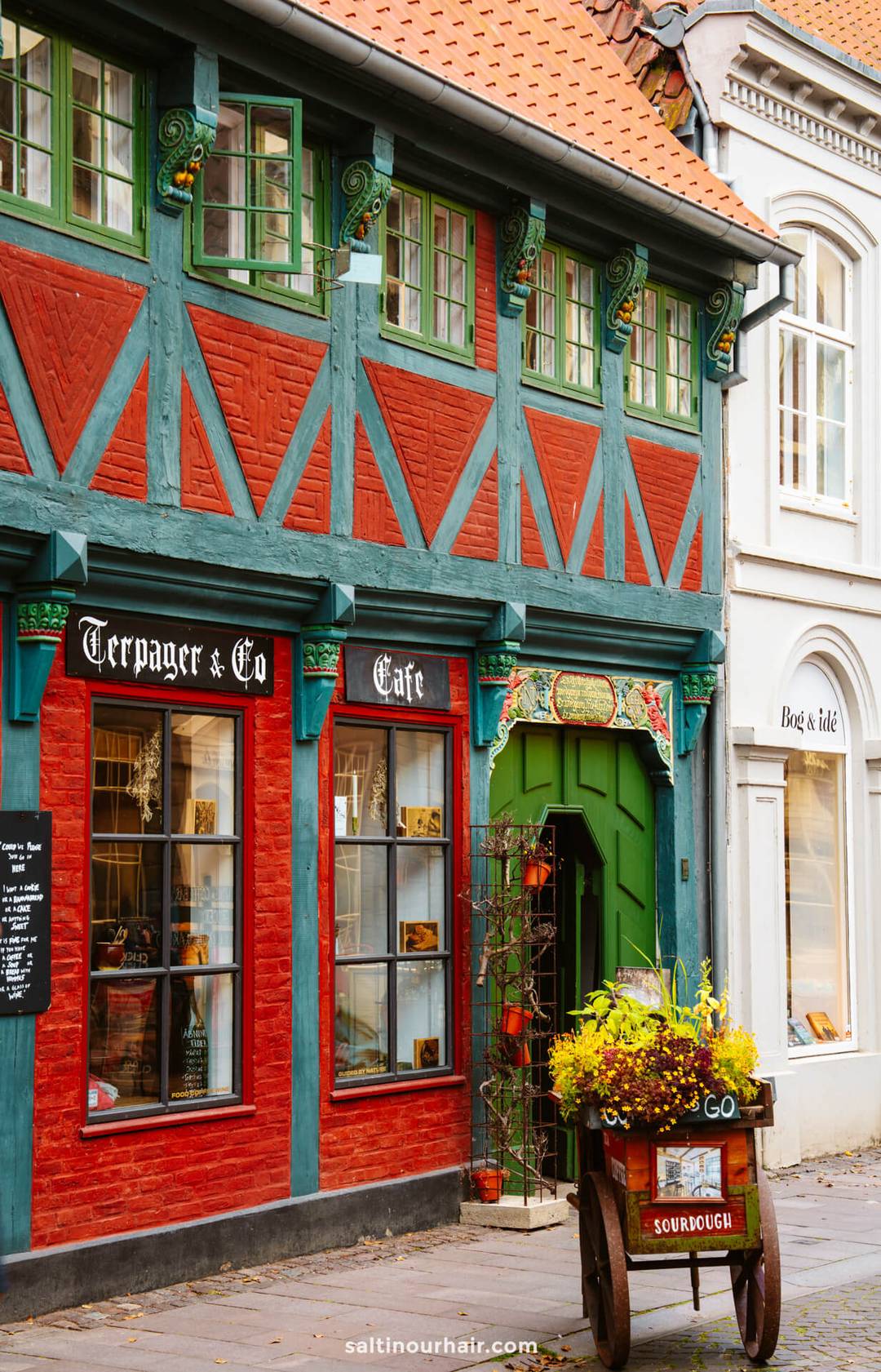 A traditional half-timbered building with teal and red accents houses a caf&eacute; and shop in Ribe; perfect for your Denmark itinerary, a cart with flowers and a &ldquo;sourdough&rdquo; sign stands outside the entrance.