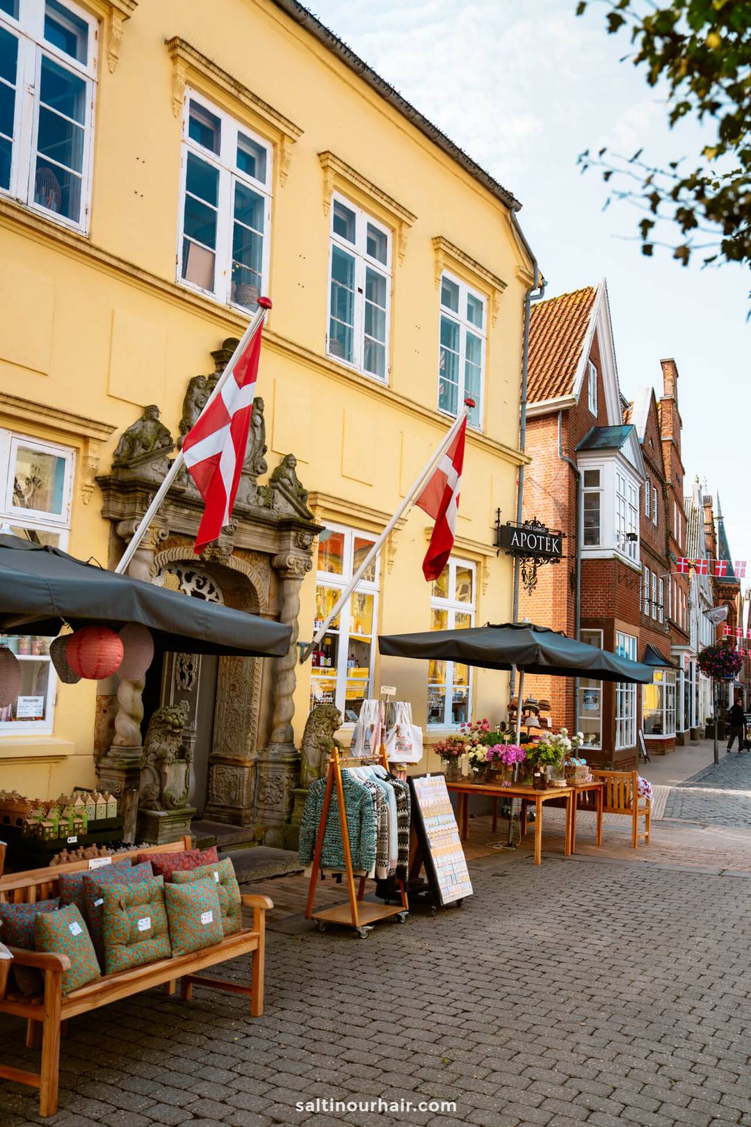 A street view of a yellow building with Danish flags in T&oslash;nder, outdoor displays of pillows and flowers, and neighboring shops&mdash;an inviting scene perfect for your Denmark itinerary in a charming European town.
