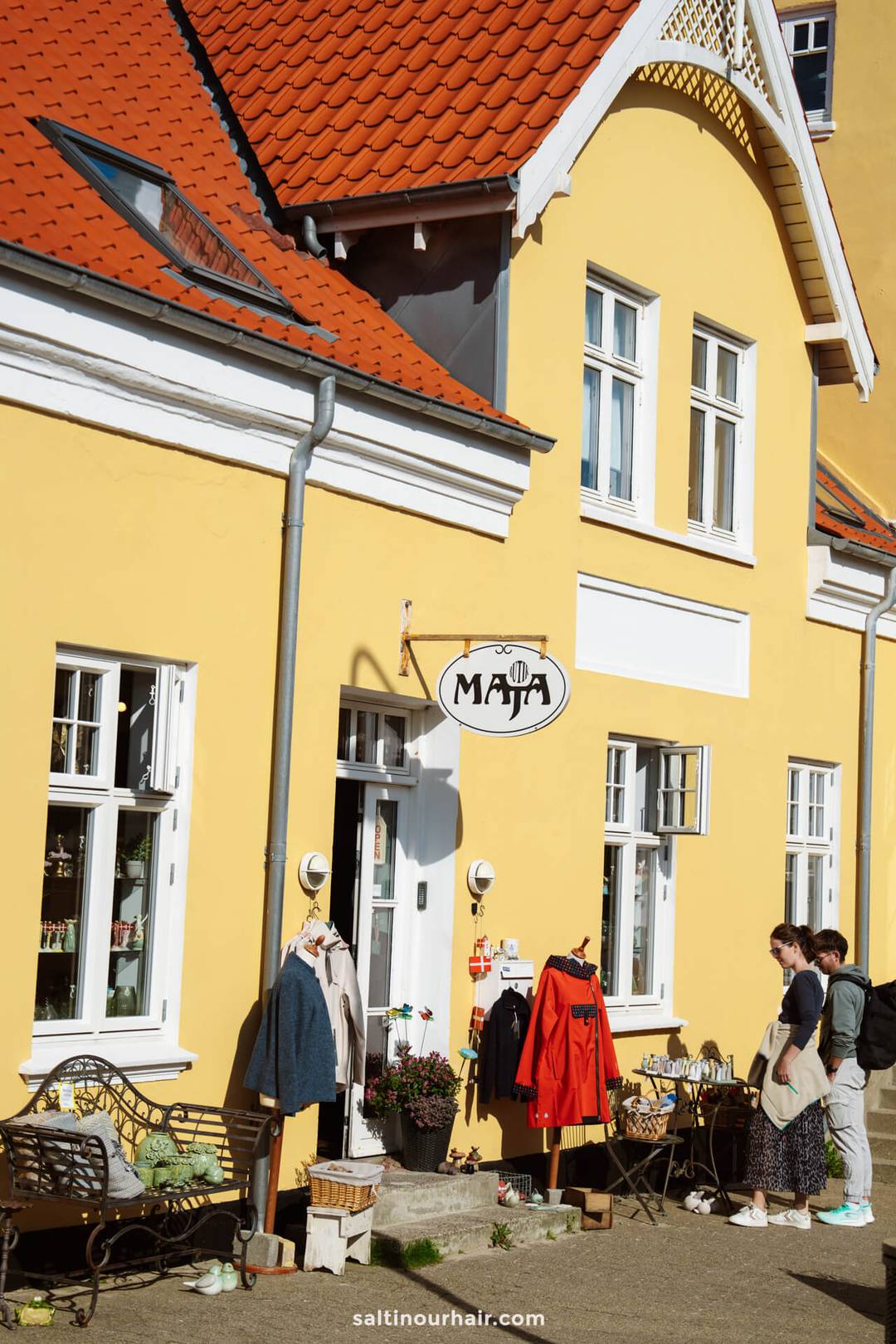 A yellow building with a red roof features a shop in L&oslash;nstrup; perfect for any Denmark itinerary, clothes and items are displayed outside, and three people stand by the entrance.