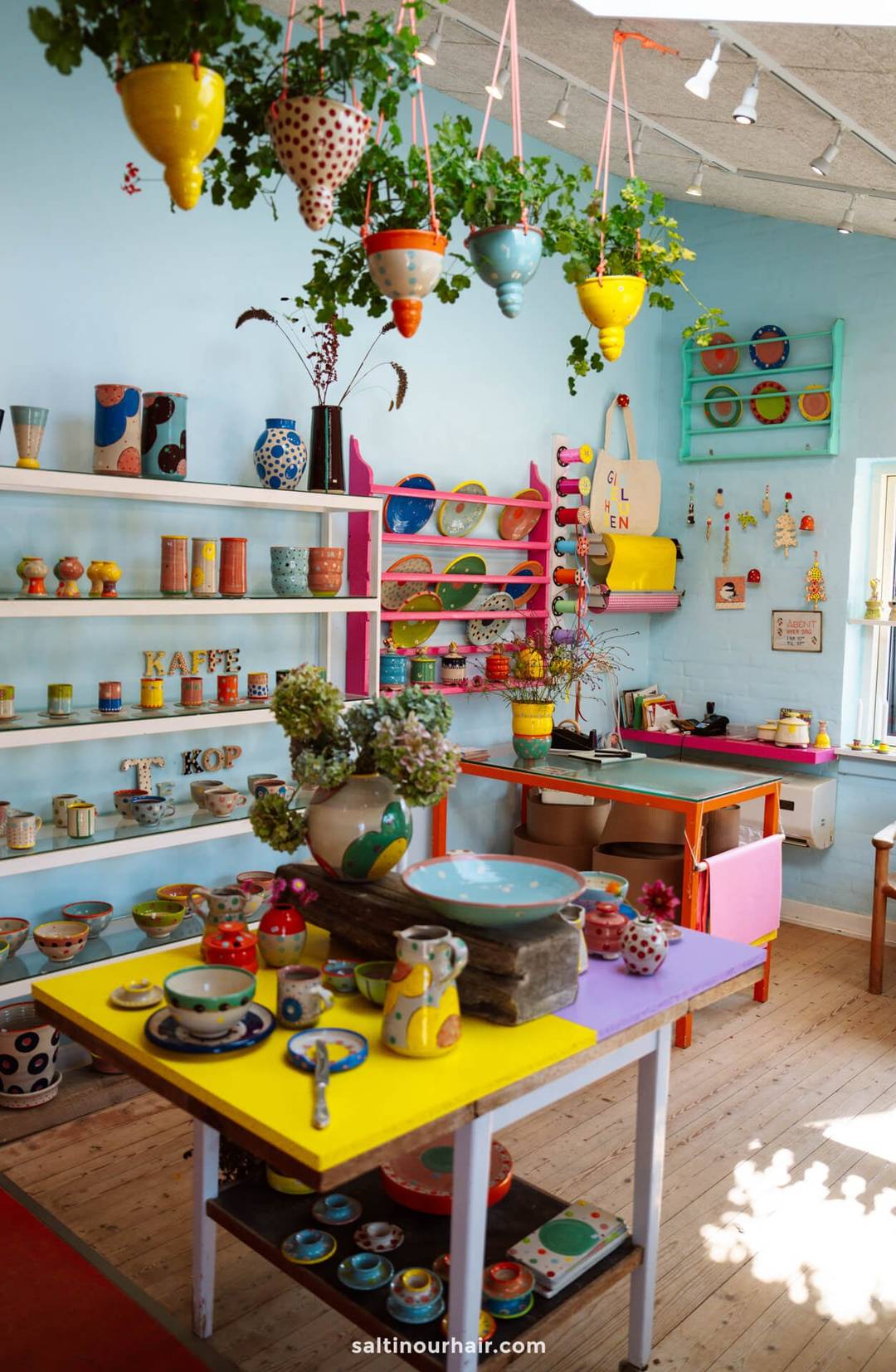 A brightly colored pottery shop in L&oslash;nstrup, perfect for any Denmark itinerary, displays various handmade ceramics&mdash;mugs, bowls, and vases&mdash;along with hanging plants and shelves against pastel blue walls.