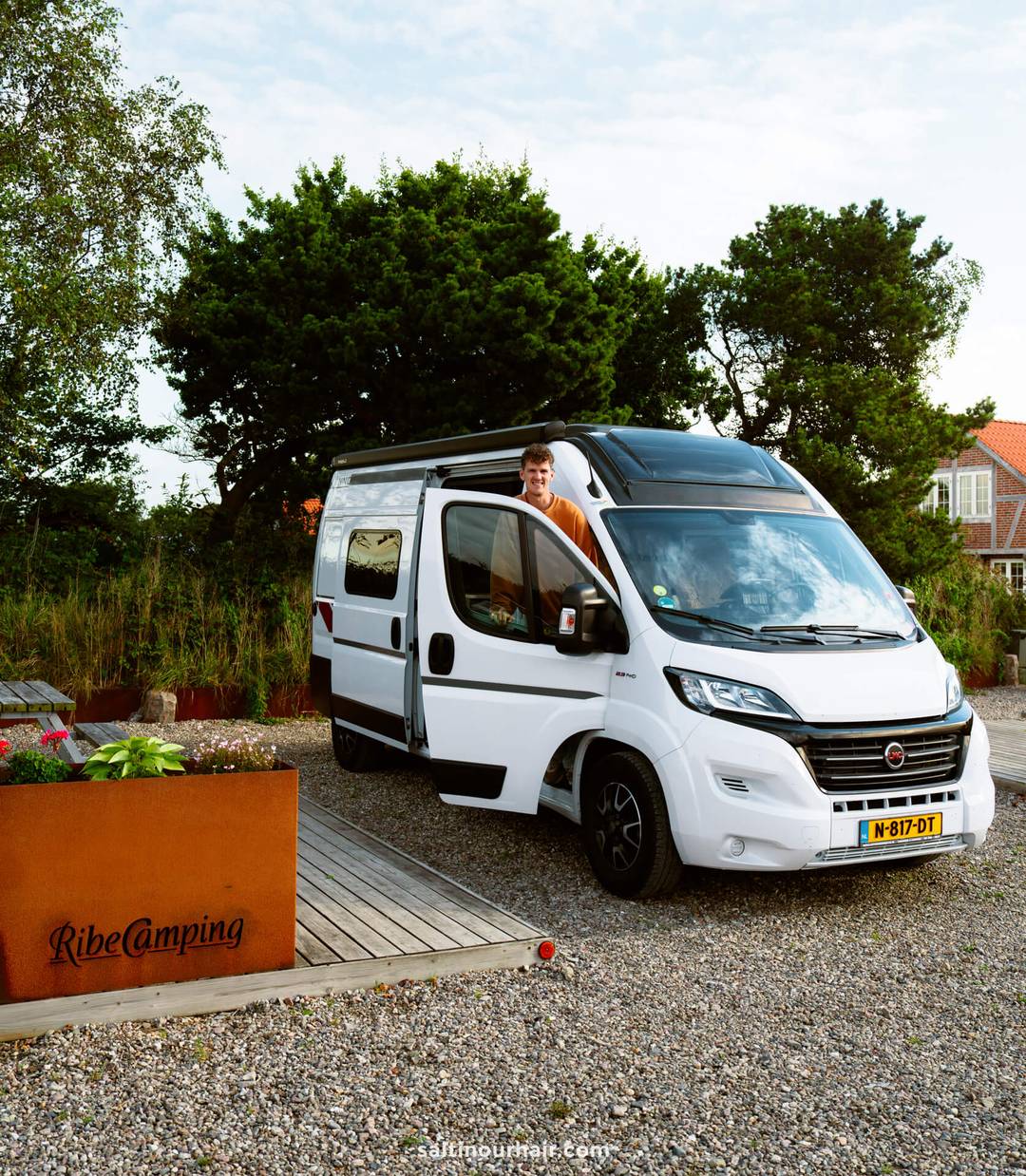 A person stands in the open side door of a white camper van parked at Ribe Camping, Denmark&mdash;a perfect stop on any Denmark road trip&mdash;with trees and buildings in the background.