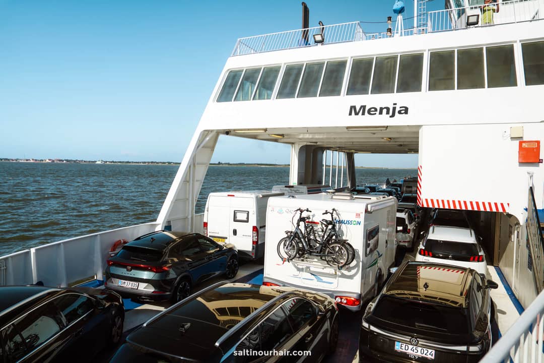 Cars and a camper van with bicycles on the back are parked on the open deck of a ferry to Fan&oslash; island, an ideal sight for any Denmark itinerary, as it travels across serene waters under a clear sky.