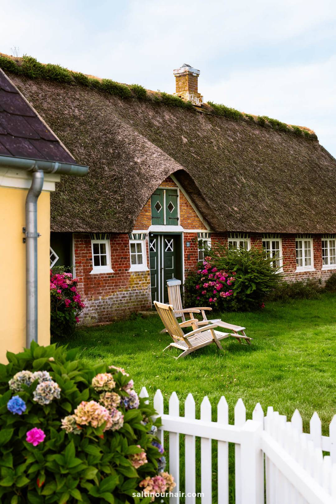 A traditional thatched-roof brick house with a green door in S&oslash;nderho, perfect for your Denmark itinerary, is surrounded by flowers and a white picket fence, with two wooden chairs inviting you to relax on the lawn.