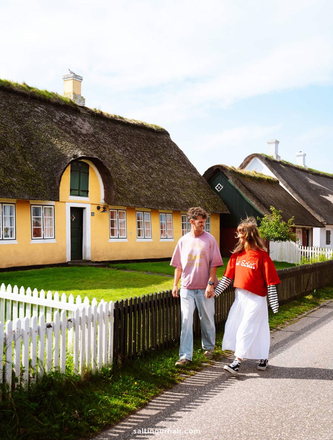 Two people walk on a road beside yellow houses with thatched roofs, white picket fences, and green lawns on a sunny day in S&oslash;nderho&mdash;an idyllic scene perfect for any Denmark itinerary.