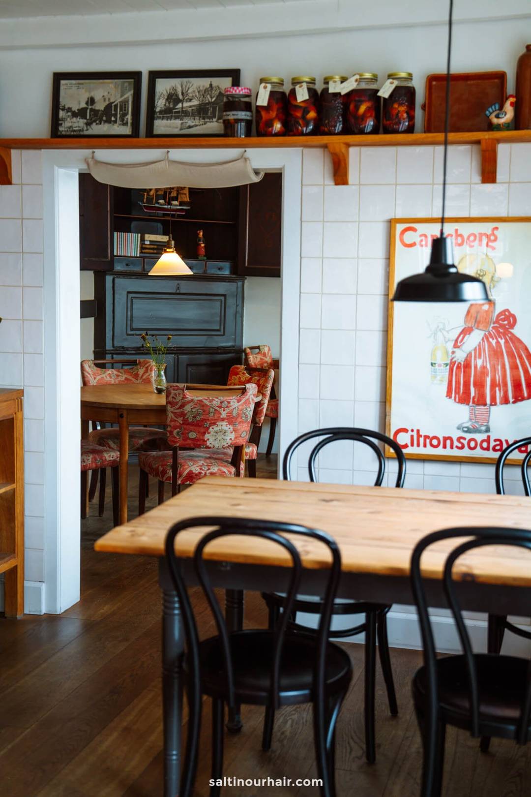 A cozy caf&eacute; interior perfect for planning your Denmark itinerary, with wooden tables and chairs, floral upholstered seating, framed art, and glass jars on a high shelf. A small vase with flowers sits on a table in the background.