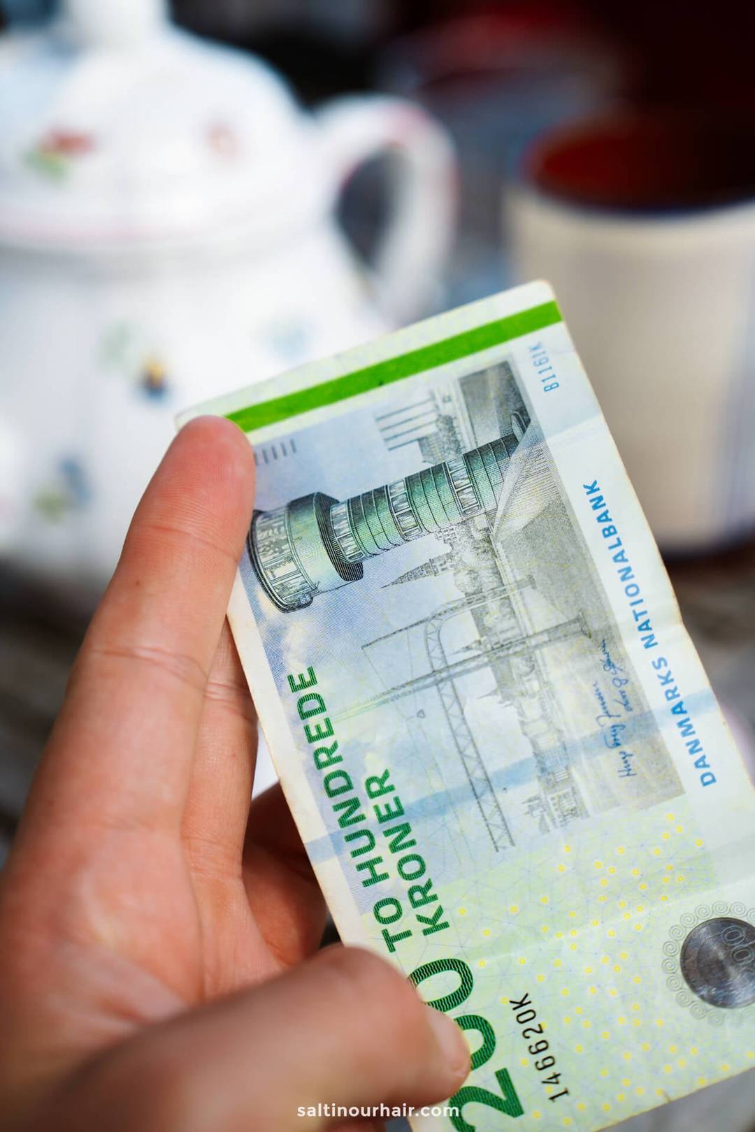 A hand holding a Danish 200 kroner banknote, with a teapot and cup blurred in the background&mdash;perfect inspiration as you plan your Denmark itinerary.