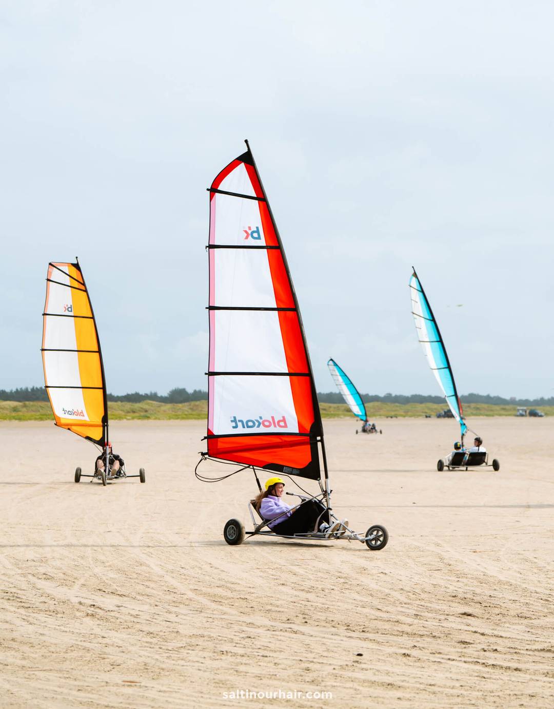 Three people ride blokarts with colorful sails on a sandy beach under a cloudy sky, with grassy dunes in the background&mdash;a perfect addition to your Denmark itinerary.