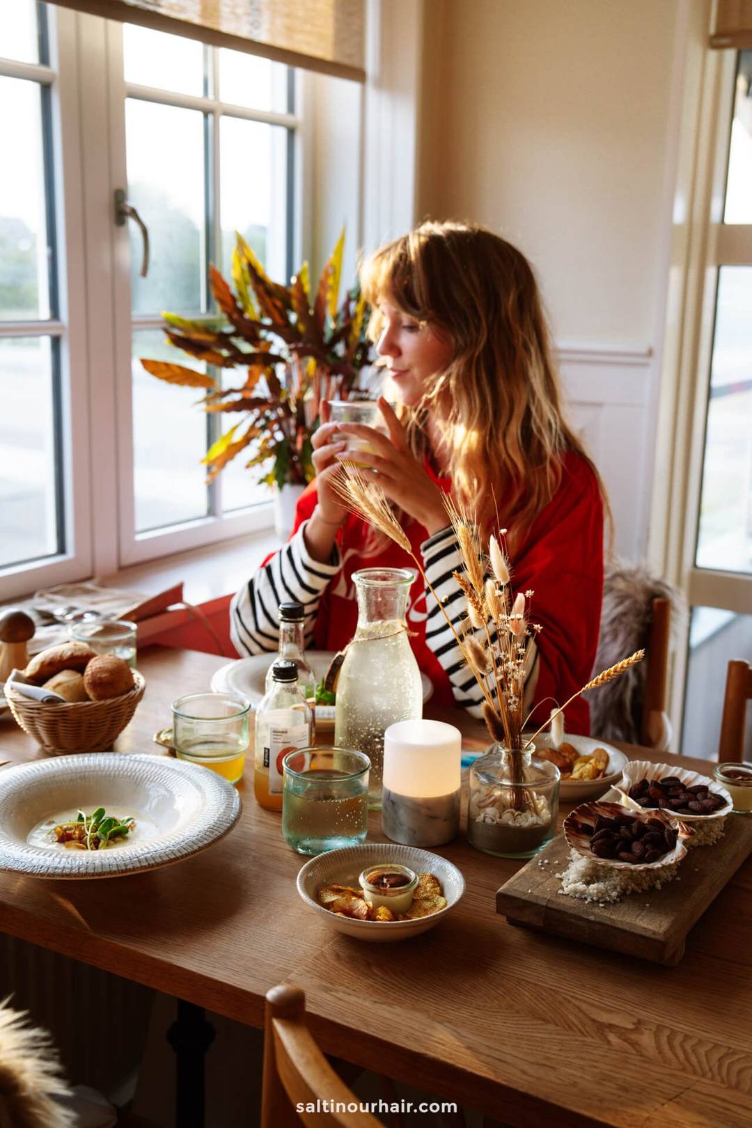 A woman sits at a wooden table set with food, drinks, and candles near a window, holding a glass and looking outside&mdash;perhaps reminiscing about her recent Denmark itinerary.