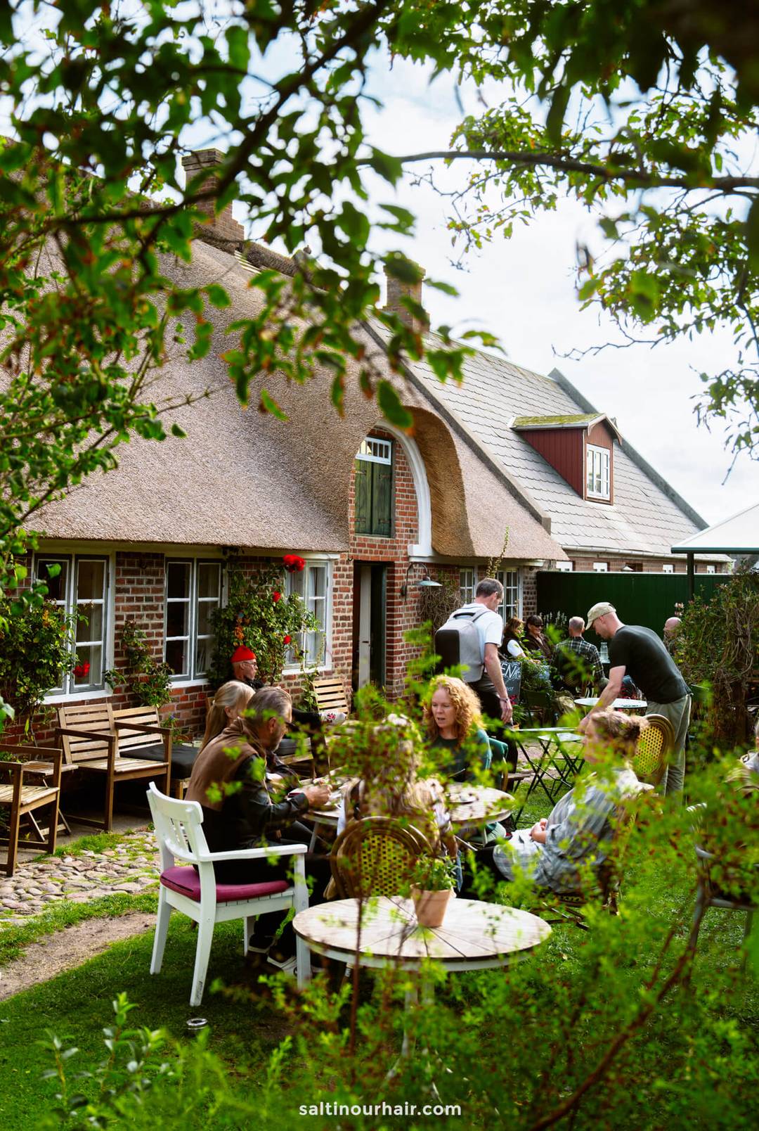 People are sitting at outdoor tables eating and talking in a garden beside a brick house with a thatched roof&mdash;a charming stop on any Denmark itinerary&mdash;enjoying the sunshine and lively atmosphere.