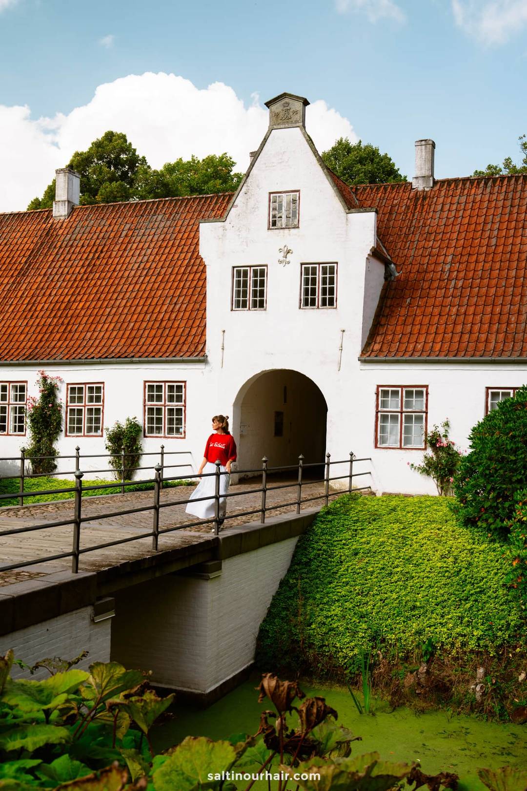 A person in a red shirt walks across a small bridge toward a white historic building with a red-tiled roof in M&oslash;gelt&oslash;nder&mdash;an ideal stop on any Denmark itinerary.