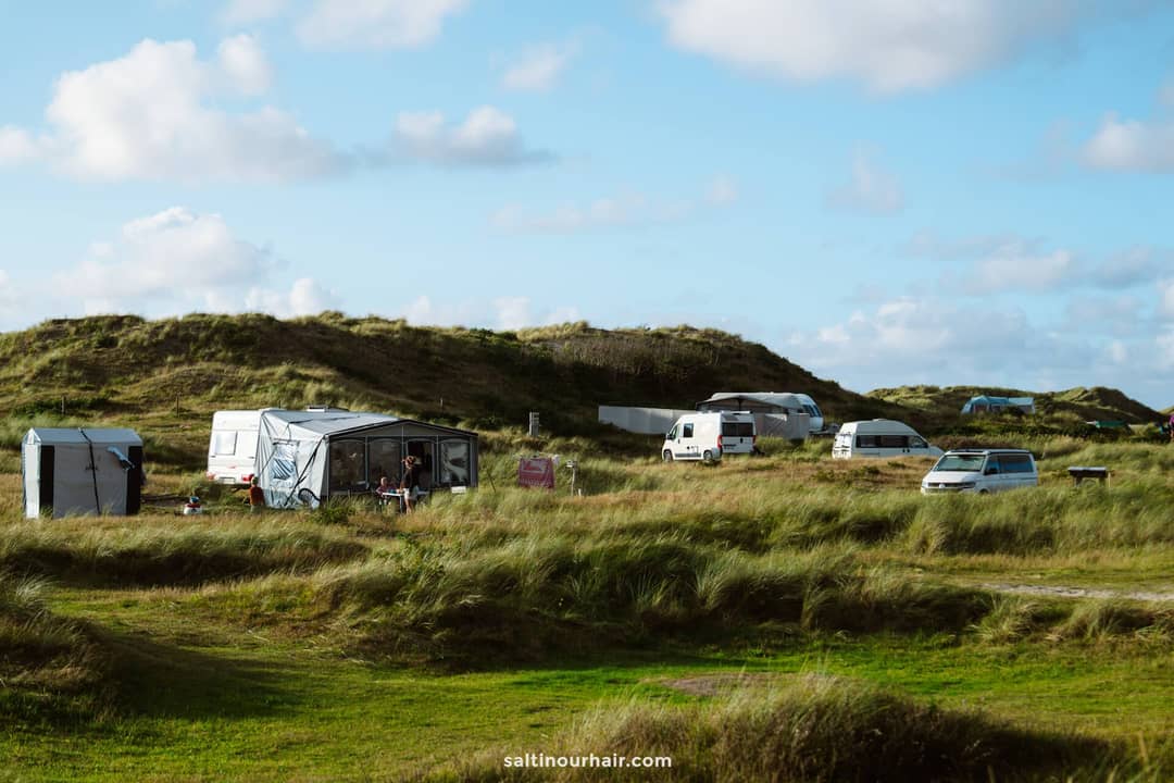 Several camper vans, tents, and cars are parked among grassy sand dunes under a partly cloudy sky on a camping site&mdash;an ideal stop on any Denmark itinerary.