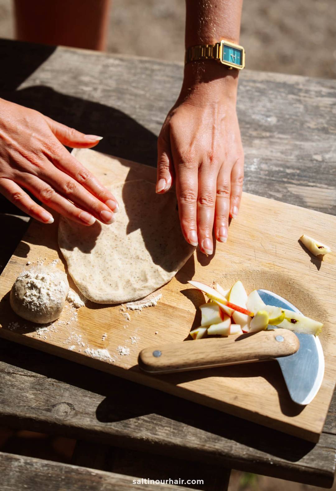 Two hands flatten dough on a wooden cutting board next to a ball of dough, sliced apples, a dough cutter, cheese pieces&mdash;perfect inspiration for planning recipes into your Denmark itinerary.