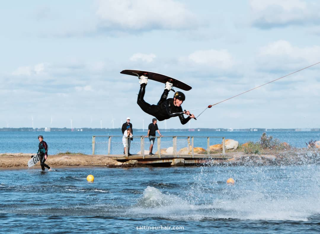 A person in a wetsuit performs an airborne trick on a wakeboard, holding a tow rope, while spectators&mdash;perhaps planning their Denmark itinerary&mdash;watch from the water&rsquo;s edge.