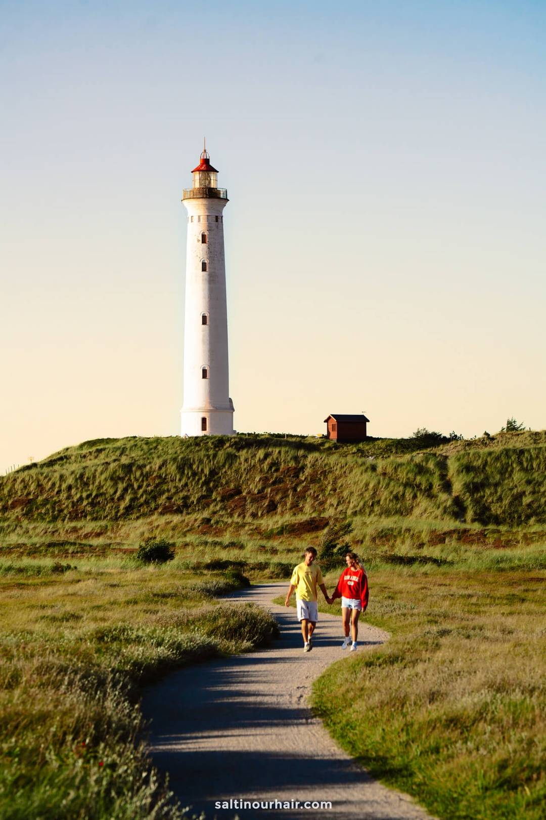 Two people walk on a path through grassy fields toward Lyngvig Fyr, Denmark&rsquo;s tall white lighthouse with a red top, under a clear sky.