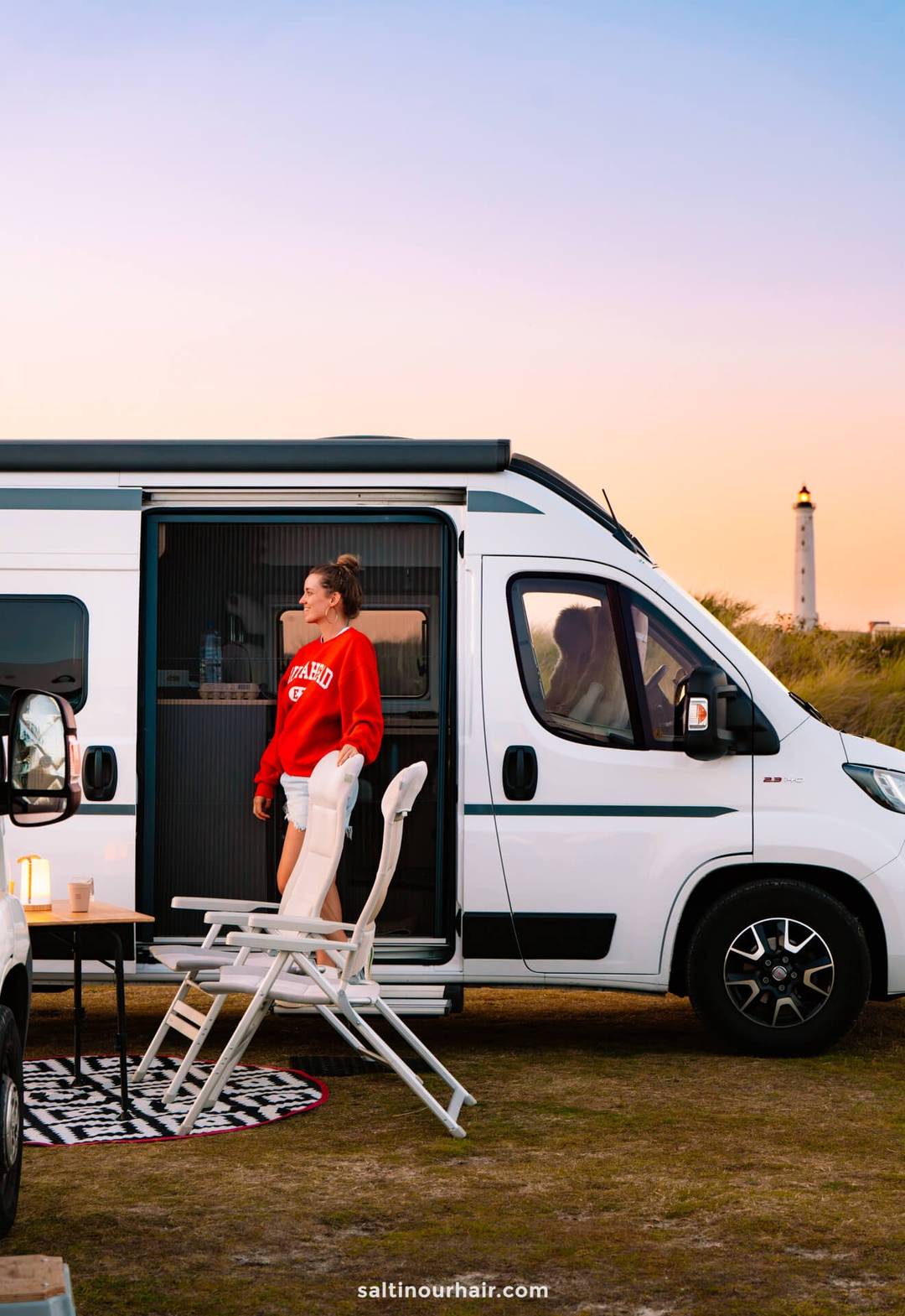 A woman stands in the doorway of a white camper van parked on grass at sunset, with a chair, table, and Lyngvig Fyr lighthouse in the background&mdash;an idyllic stop on any Denmark road trip.