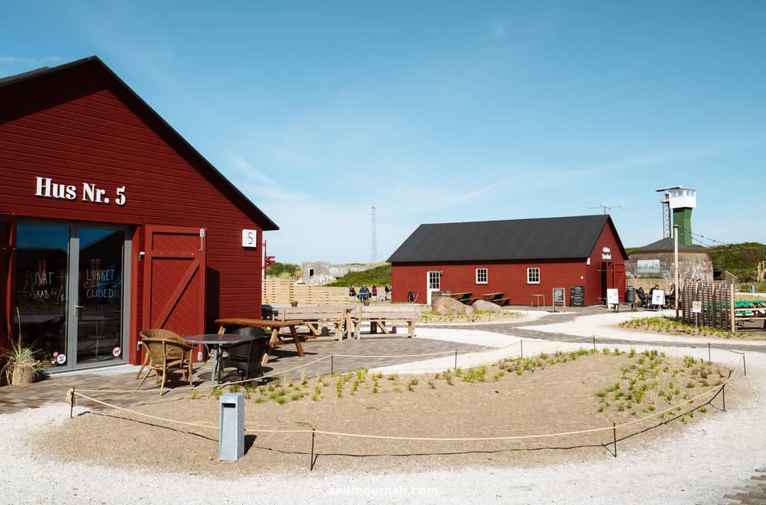 Scene with red wooden buildings, outdoor seating, gravel paths, and sparse landscaping under a clear blue sky&mdash;a charming stop to include on your Denmark itinerary. A watchtower rises in the background.
