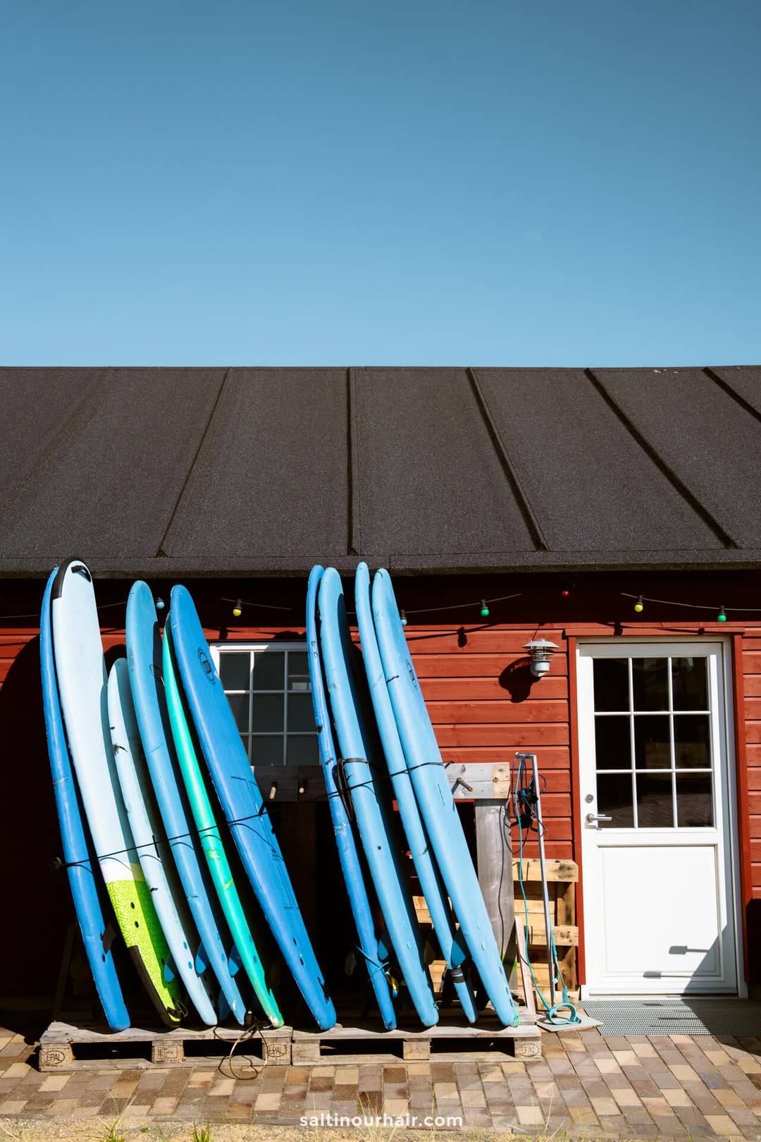 Several surfboards are propped against a red wooden building with a black roof, next to a white door and outdoor string lights&mdash;an inviting spot for any Denmark road trip itinerary under a clear blue sky.