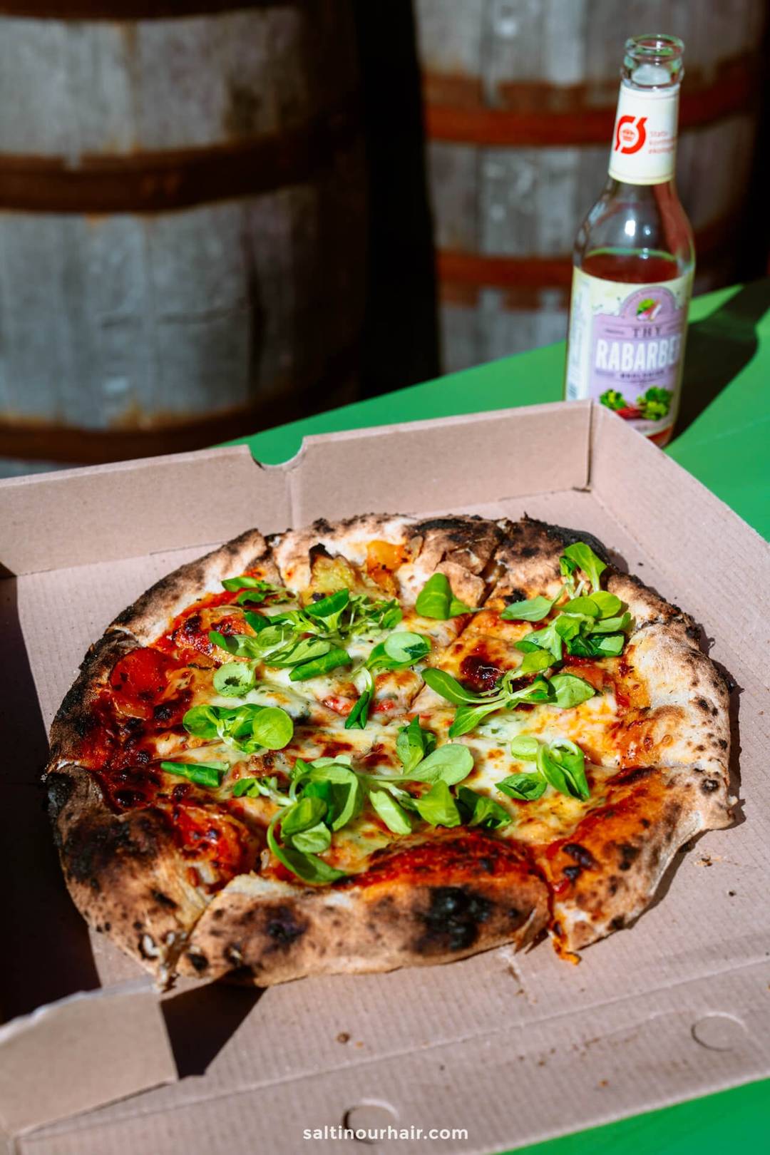 A wood-fired pizza topped with green leaves sits in an open cardboard box on a green table next to a bottle of rhubarb soda&mdash;perfect for your Denmark itinerary. Two wooden barrels are visible in the background.
