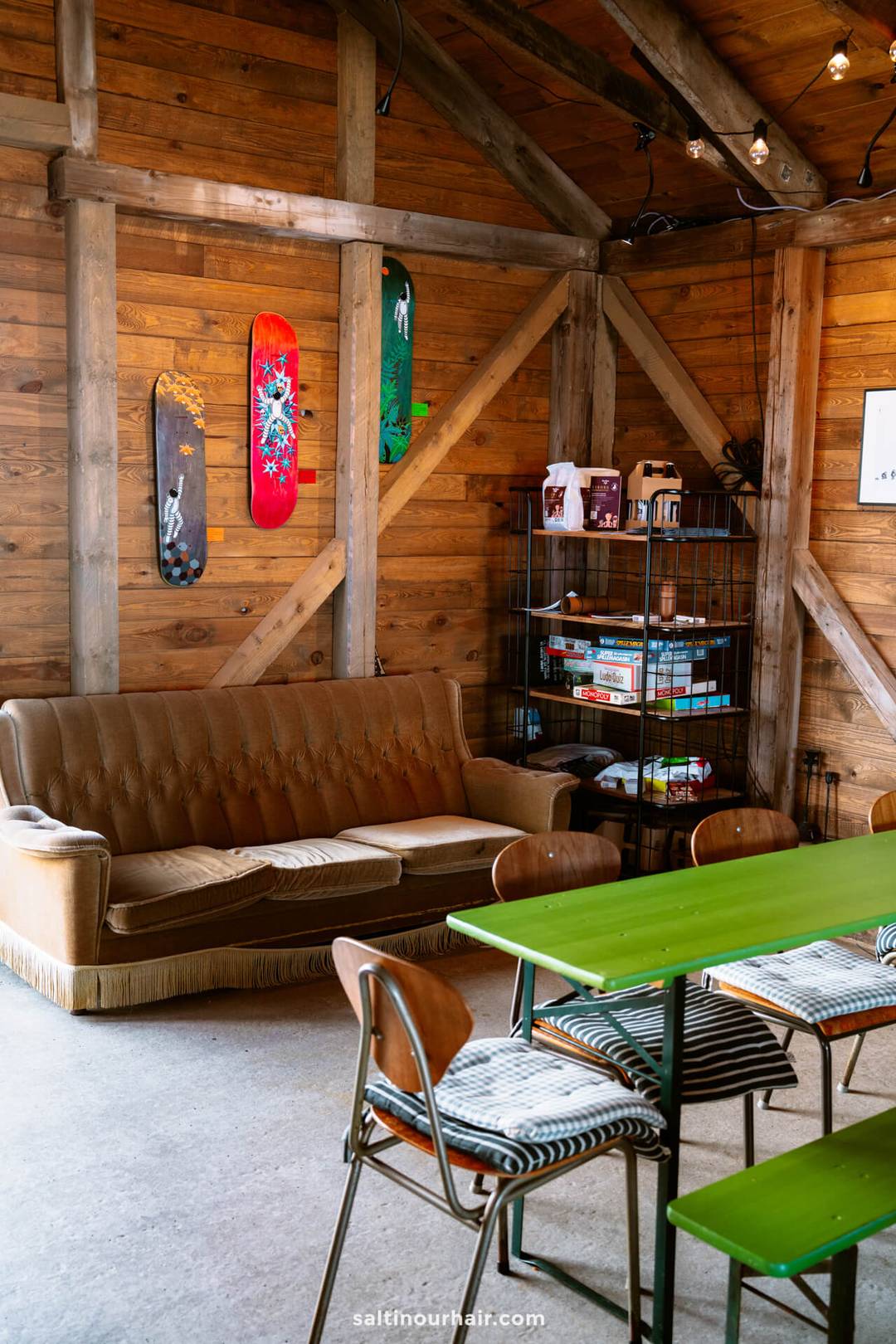 A cozy room with a brown sofa, green tables, wooden chairs, and a metal shelf of board games&mdash;perfect for planning your Denmark itinerary among skateboards mounted on wooden walls.