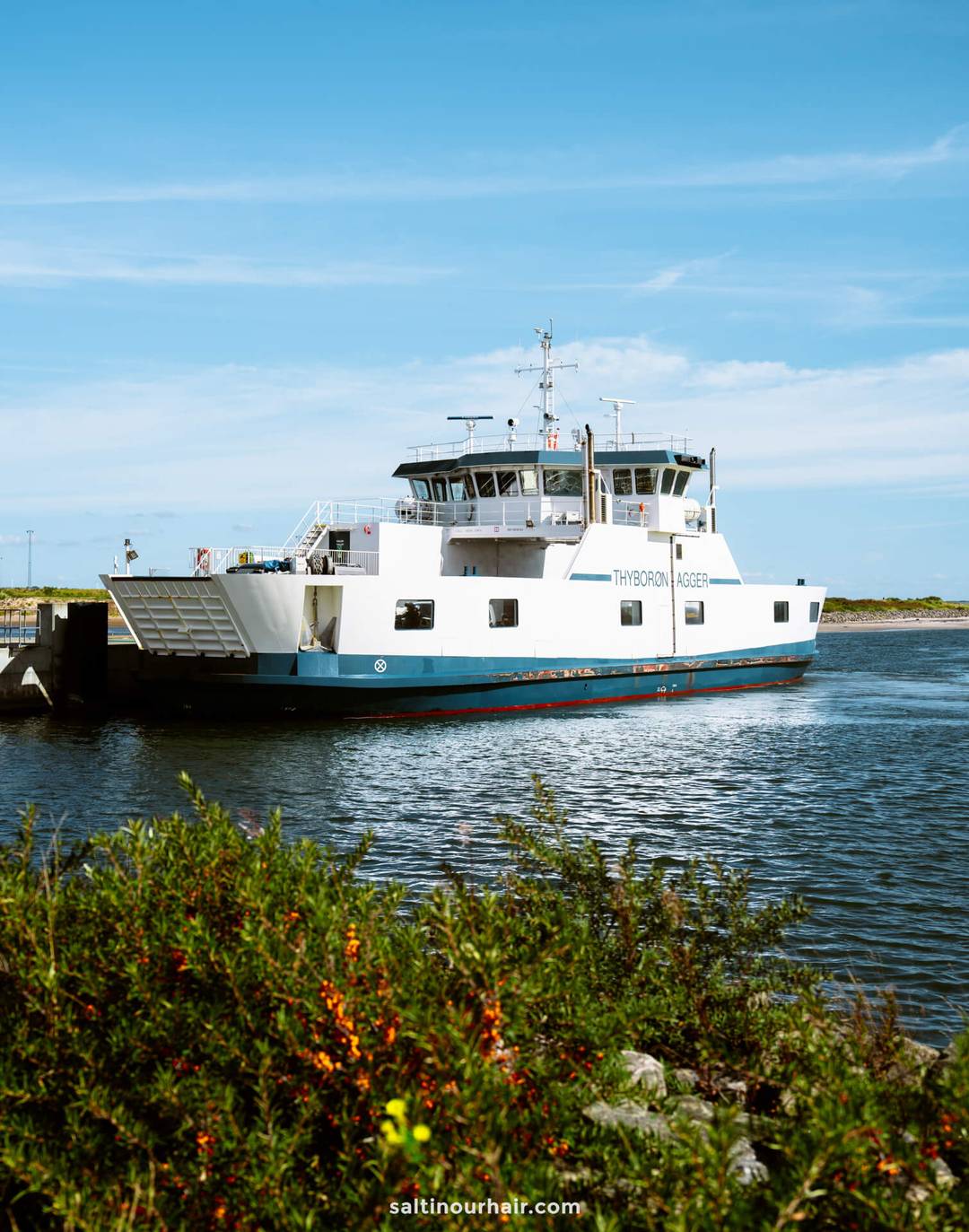 A white and blue ferry named Thybor&oslash;n-Agger is docked near the shore with greenery and water in the foreground under a clear sky, making it a scenic stop on any Denmark itinerary.