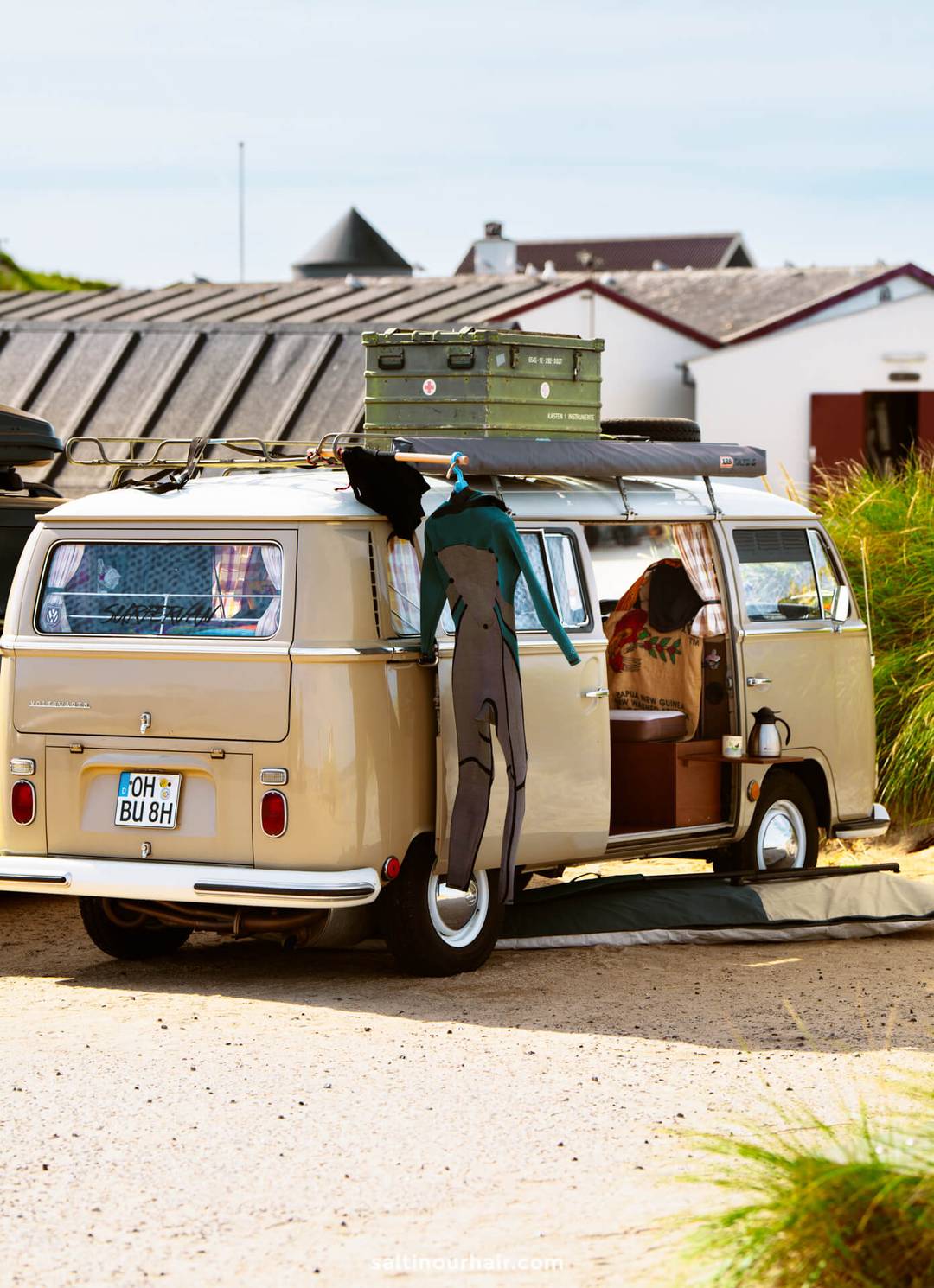 A beige camper van with surf gear, including a wetsuit hanging on the side, parked near beach houses on a sunny day&mdash;perfect for any Denmark itinerary.