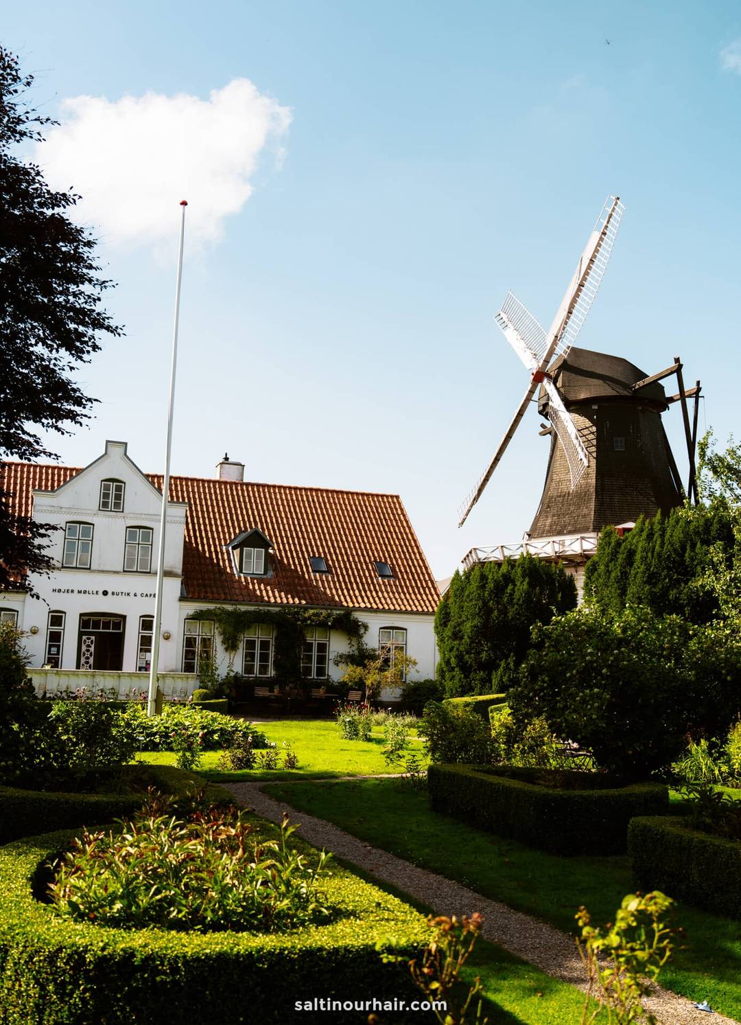 A traditional Dutch windmill stands next to a white house with a red-tiled roof in H&oslash;jer, surrounded by a manicured garden under a blue sky&mdash;an idyllic scene to inspire any Denmark itinerary.