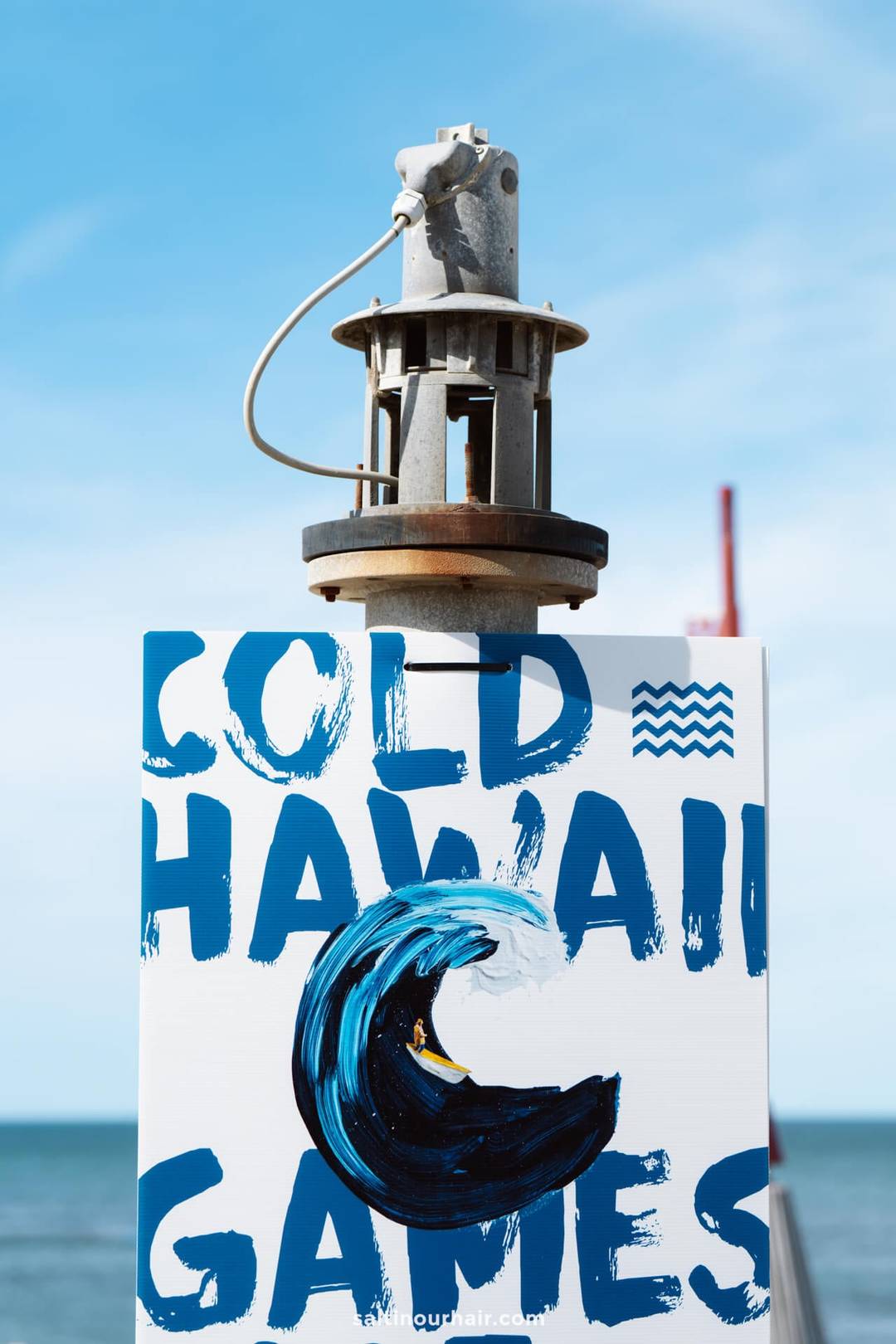 A sign reading Cold Hawaii Games with a painted wave sits beneath mechanical equipment, set against a blue sky and ocean&mdash;an ideal stop on any Denmark itinerary.