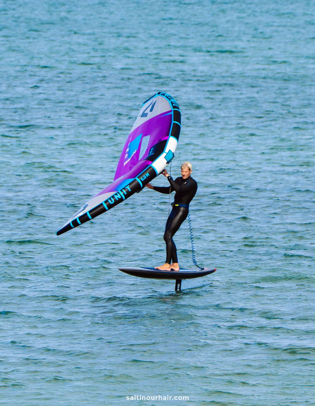 A person wearing a wetsuit rides a hydrofoil board on the water, holding a purple and blue handheld sail&mdash;an adventure you might add to your Denmark itinerary.