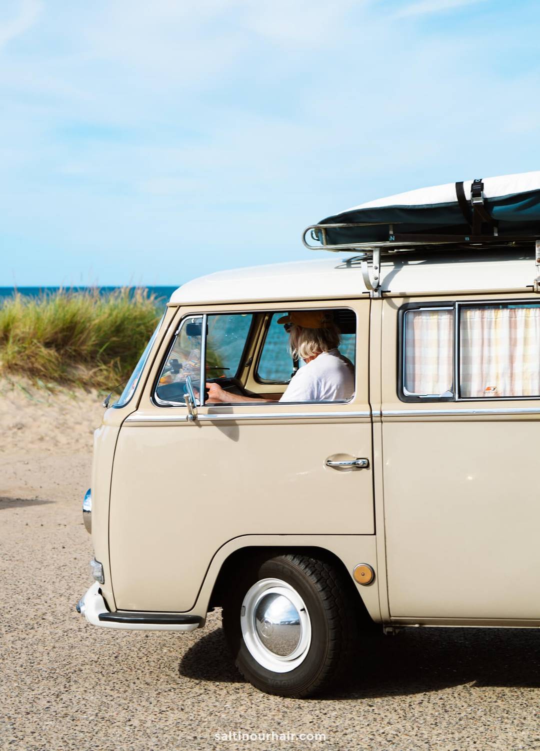 A person drives a beige vintage van with a roof rack near a sandy beach and grassy dunes under a blue sky, capturing the spirit of a classic Denmark road trip.
