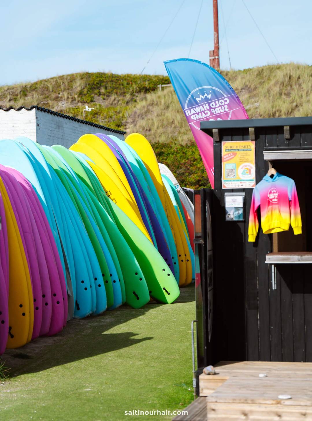Colorful surfboards are lined up next to a black wooden hut with a rainbow sweatshirt hanging outside&mdash;an inviting stop on any Denmark itinerary, with a flag and grassy sand dunes visible in the background.