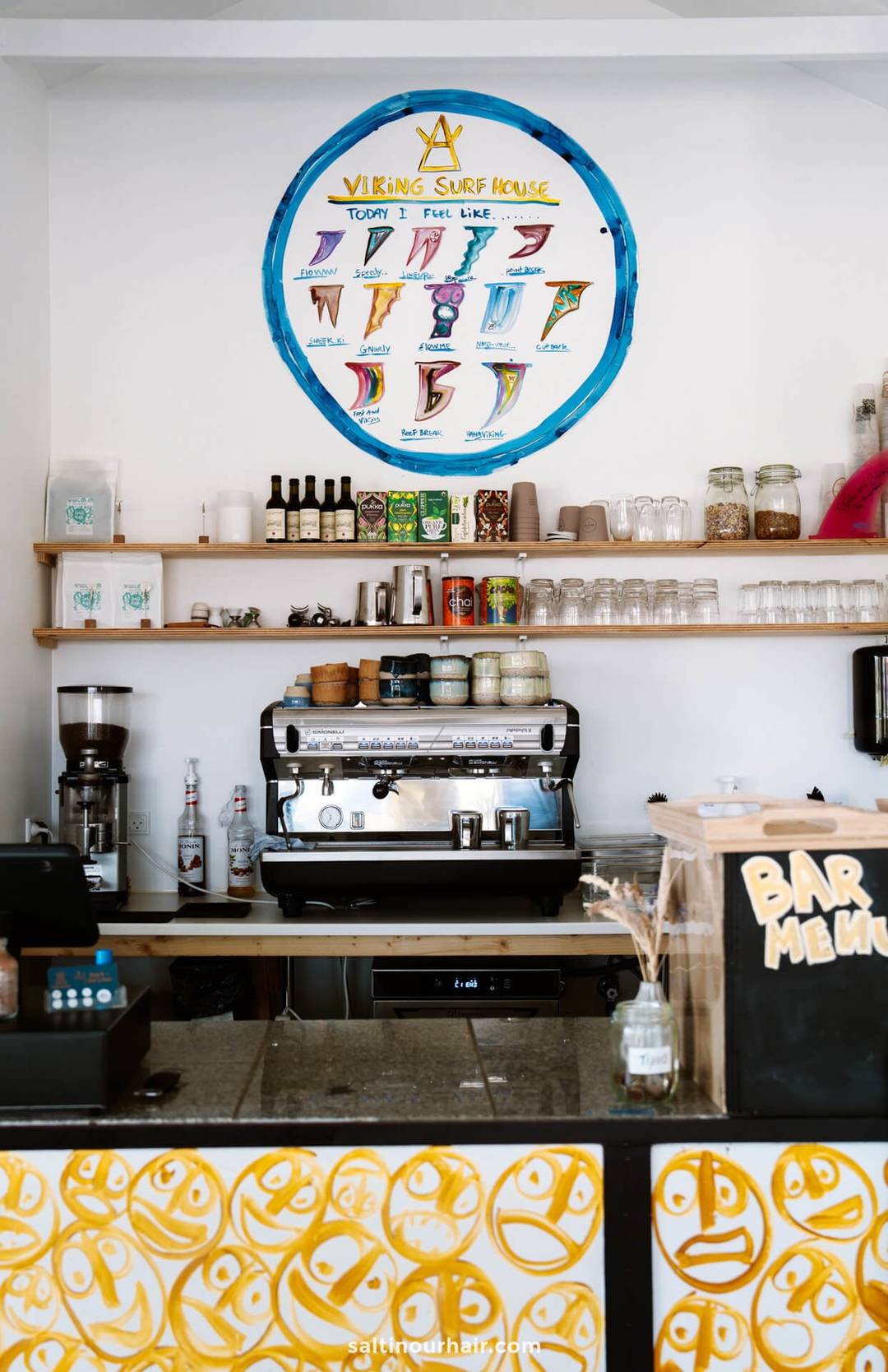 A coffee bar with a large espresso machine, shelves of cups and jars, and a colorful surf-themed menu displayed on the wall above&mdash;an inviting stop to add to your Denmark itinerary.