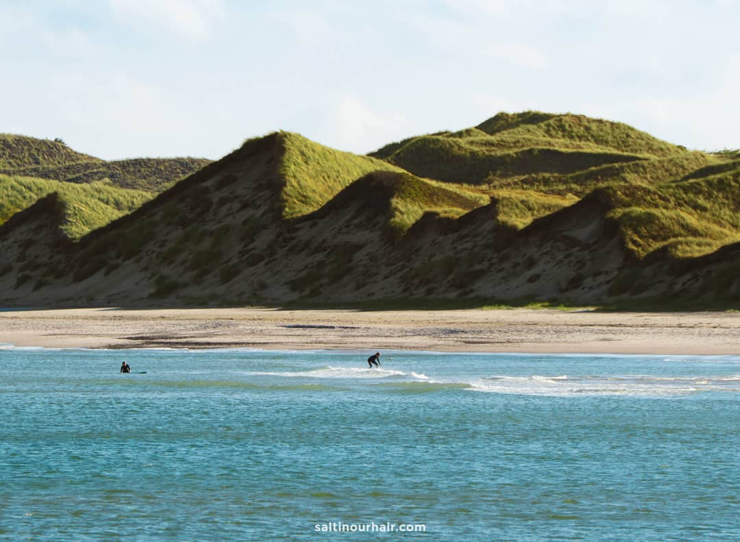 Two people surf small waves near a sandy beach with grassy, sloping dunes in the background under a partly cloudy sky&mdash;a perfect scene to include in your Denmark itinerary.