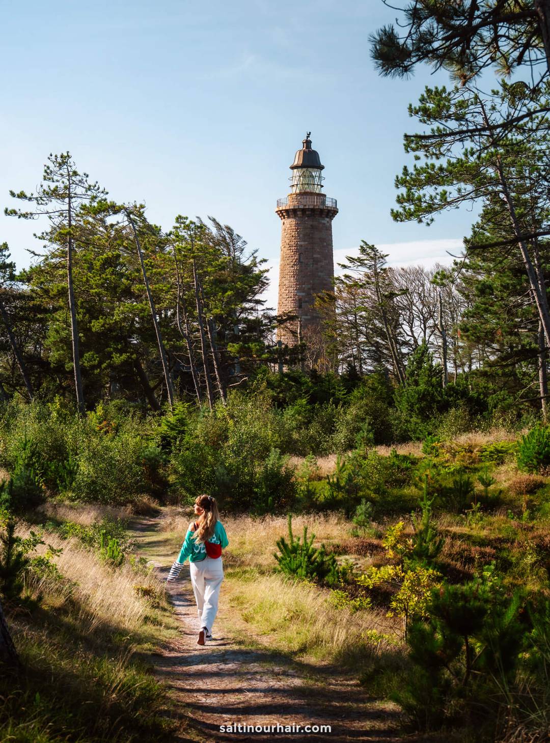 A person walks along a forest path toward the tall brick lighthouse Lodbjerg Fyr surrounded by trees on a sunny day&mdash;a perfect stop on any Denmark itinerary.