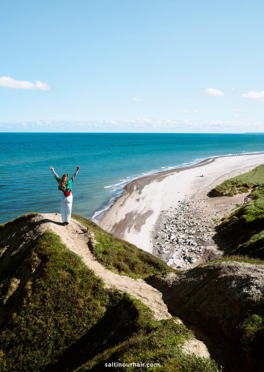 A person stands on a grassy cliff called Bulbjerg Klint overlooking a sandy beach and blue ocean under a clear sky, arms raised in the air&mdash;an inspiring stop on any Denmark itinerary.