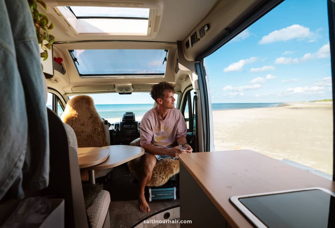 A man sits inside a camper van parked by a sandy beach, looking out at the ocean through the open side door under a partly cloudy sky&mdash;an idyllic stop on his Denmark road trip itinerary.