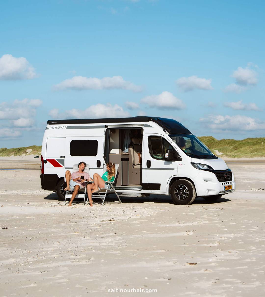 Two people sit on chairs outside a white camper van parked on a sandy beach, enjoying a peaceful stop along their Denmark road trip itinerary, with grassy dunes in the background under a blue sky with clouds.