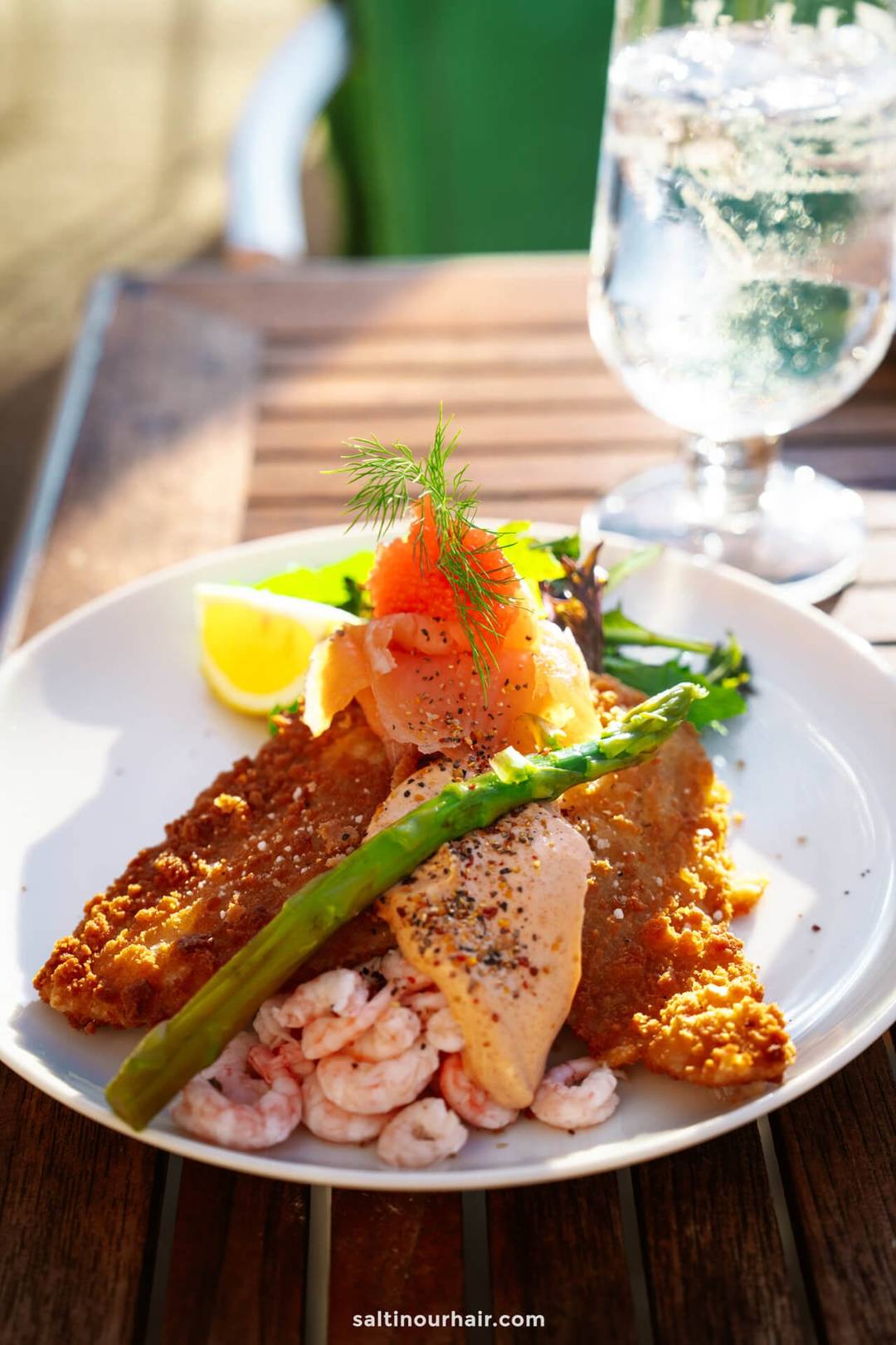 A plate of breaded fish fillets topped with smoked salmon, shrimp, a lemon wedge, asparagus, herbs, and black pepper&mdash;this Danish-inspired meal could be the highlight of your Denmark itinerary, enjoyed with a glass of water on a wooden table.