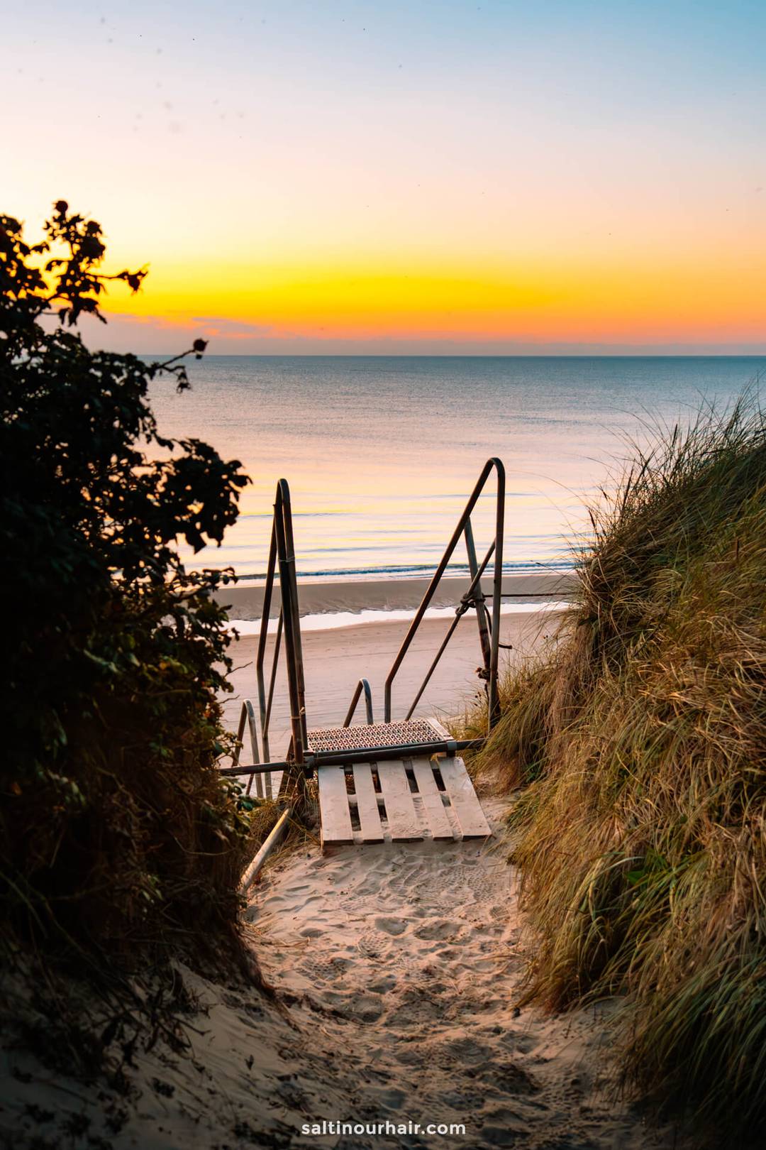 A metal staircase leads down sandy dunes to a calm beach at sunset, with orange and blue hues in the sky and gentle waves&mdash;an ideal stop on any Denmark itinerary.