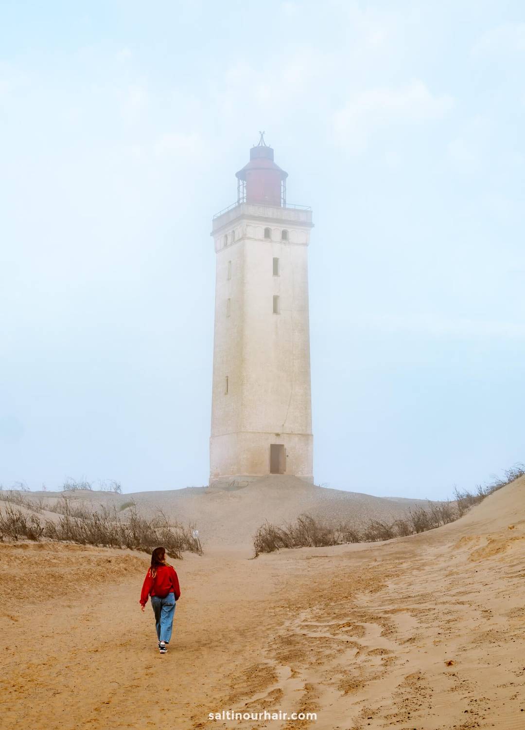 A woman in a red jacket walks toward a tall, beige lighthouse called Rubjerg Knude surrounded by sand and sparse vegetation on a foggy day&mdash;a classic scene for any Denmark itinerary.